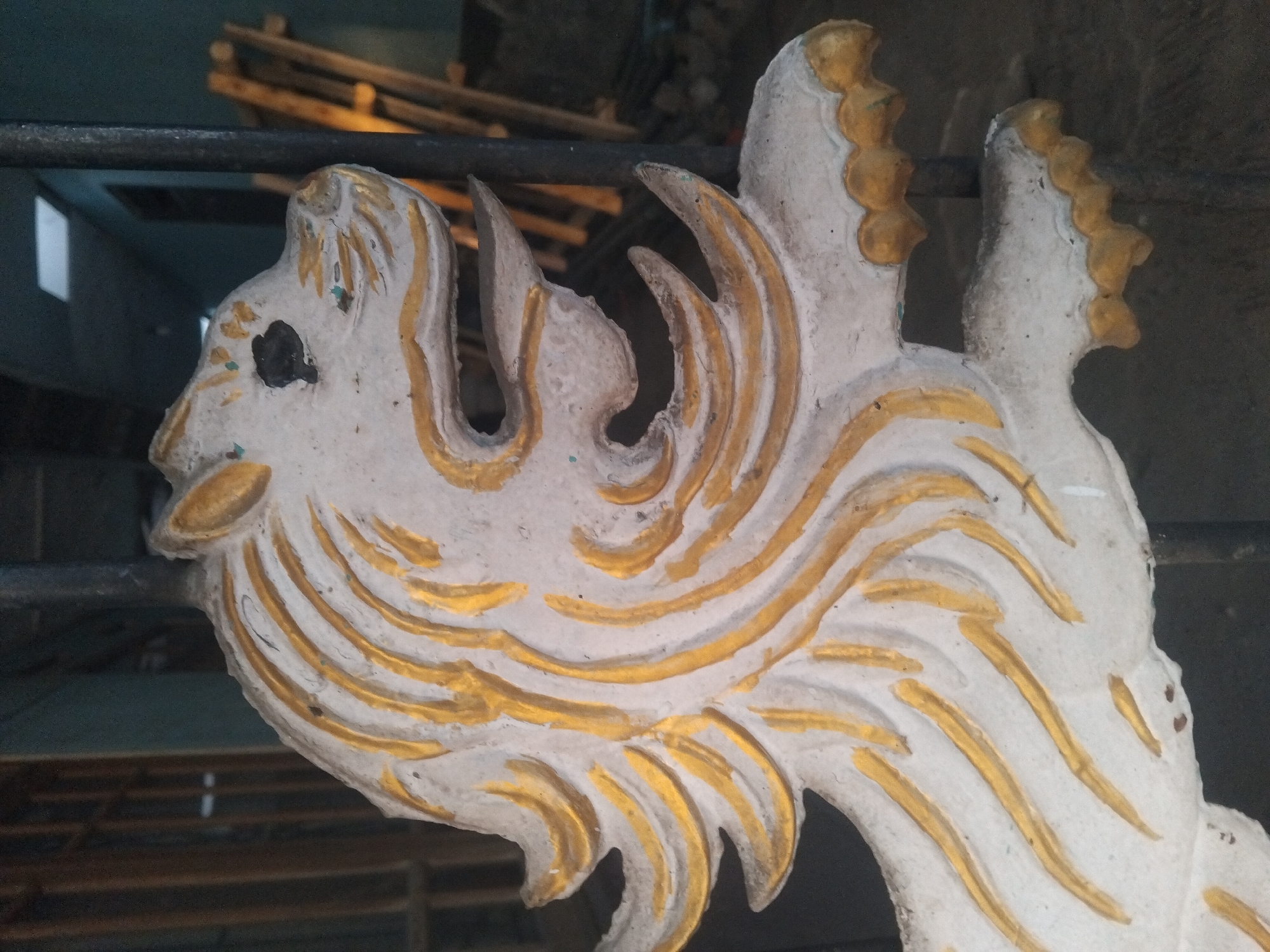 The image shows a close-up, side-view of a decorative plaster or cement lion's head with gold accents, possibly a carving or part of a larger structure. The lion's mane is depicted with flowing gold lines against a white background. The lion's mouth is open, revealing a carved opening and sharp-looking teeth. The eyes are black indentations. The lion appears to be attached to or situated in front of a dark metal bar or fence. In the background, out of focus, there are wooden structures resembling scaffolding or ladders, and hints of indoor lighting from a ceiling. The overall scene suggests an outdoor or semi-outdoor setting, possibly in a construction area or a place with decorative architectural elements. The lion's head is weathered, with some chipping and dirt visible. The quality of the image is low, with visible noise and a slightly blurred background. The location is identified as Tempeh, Indonesia.