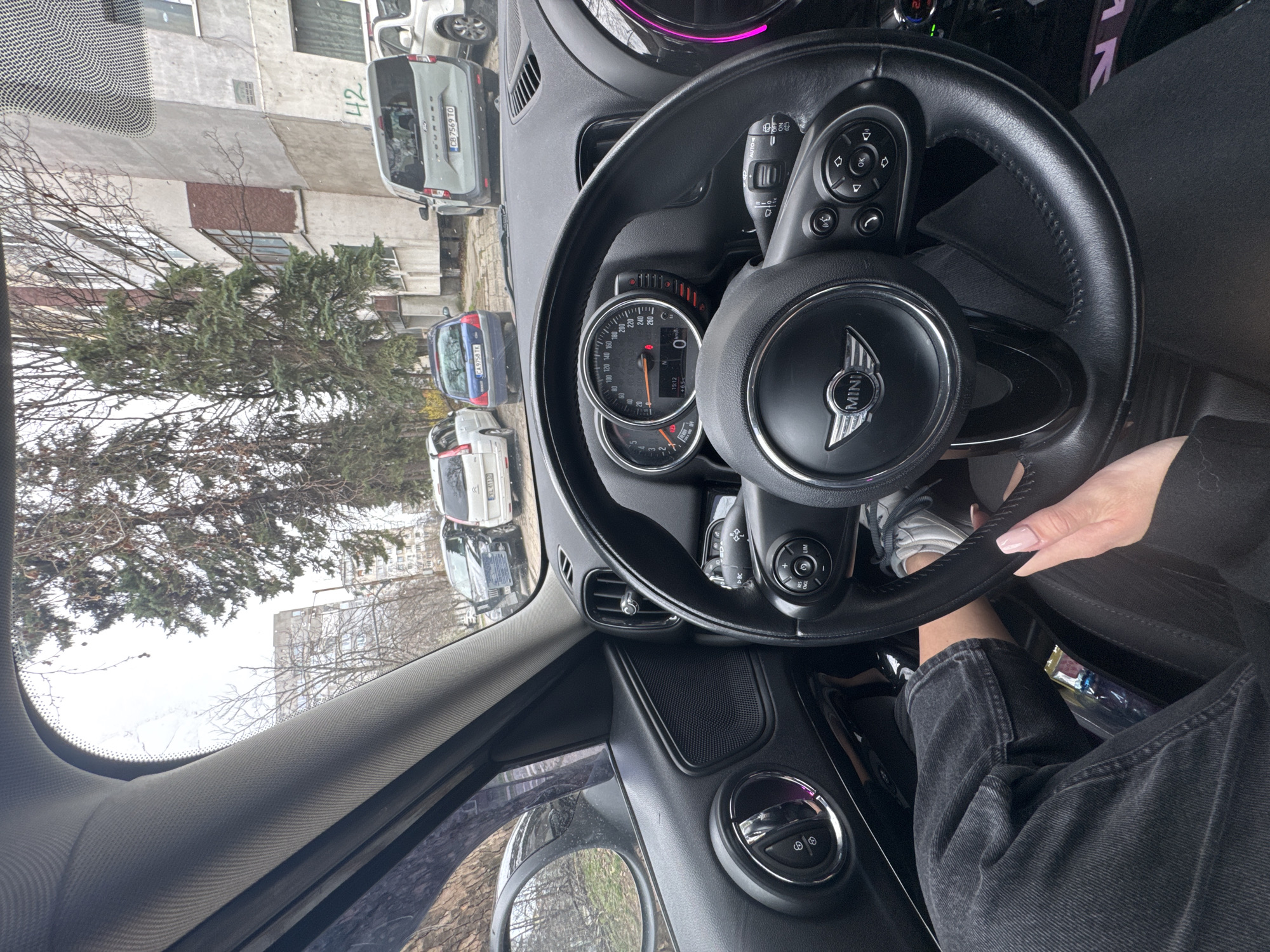 Here is a detailed description of the image:
**Setting and Environment:**
The image appears to be taken from inside a Mini Cooper, possibly in a parked position, looking out at a street scene. The location is plausibly in Sofia, Bulgaria, based on the visible license plates of the vehicles outside. The weather appears to be overcast. The scene outside includes trees, buildings (one with the number 42 prominently displayed), and several parked vehicles.
**Objects:**
* **Car Interior:** The dominant objects are the Mini Cooper's interior elements:
* Steering wheel with the MINI emblem
* Dashboard with instrument cluster (speedometer is visible)
* Center console with various controls
* Windshield and interior trim
* **Vehicles Outside:** Several vehicles can be seen outside the car, including:
* A white van with license plate "CB 7569 TO"
* A blue car with license plate "CA 9758 TK"
* A white car with license plate "XL 4113"
* **Building:** A light-colored building with the number "42" is visible in the background.
* **Personal Items:** An unidentifiable bag appears to be inside the vehicle.
**People and Actions:**
* **Driver:** A hand can be seen holding the steering wheel. The driver appears to be female, wearing dark jeans and a light-colored sneaker.
**Interactions:**
* The driver is holding the steering wheel, suggesting they are either preparing to drive, have recently parked, or are simply stationary.