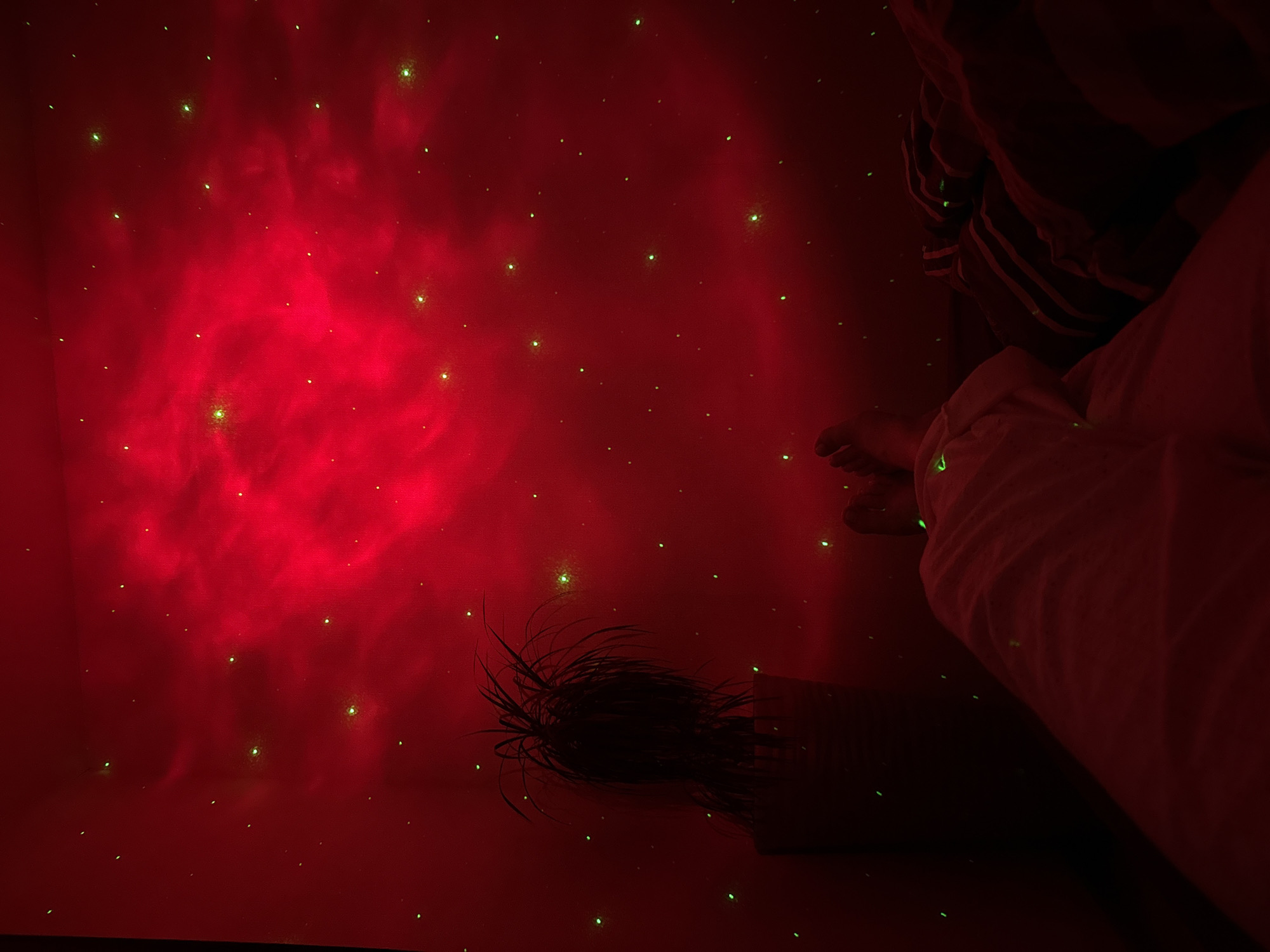 The image depicts a bedroom at night, illuminated by a red nebula projector casting colorful light and green laser stars onto the walls and ceiling. In the foreground, a person's legs are visible, clad in white pants, resting on a bed with striped bedding. To the left, a potted plant with long, thin leaves stands in a pot. The room is dark, with the only significant light source being the projector, creating an atmospheric, ethereal ambiance. The overall scene is one of relaxation and visual interest, potentially in a home environment. The location of Perm, Russia is not discernible from the visual information present in the image.