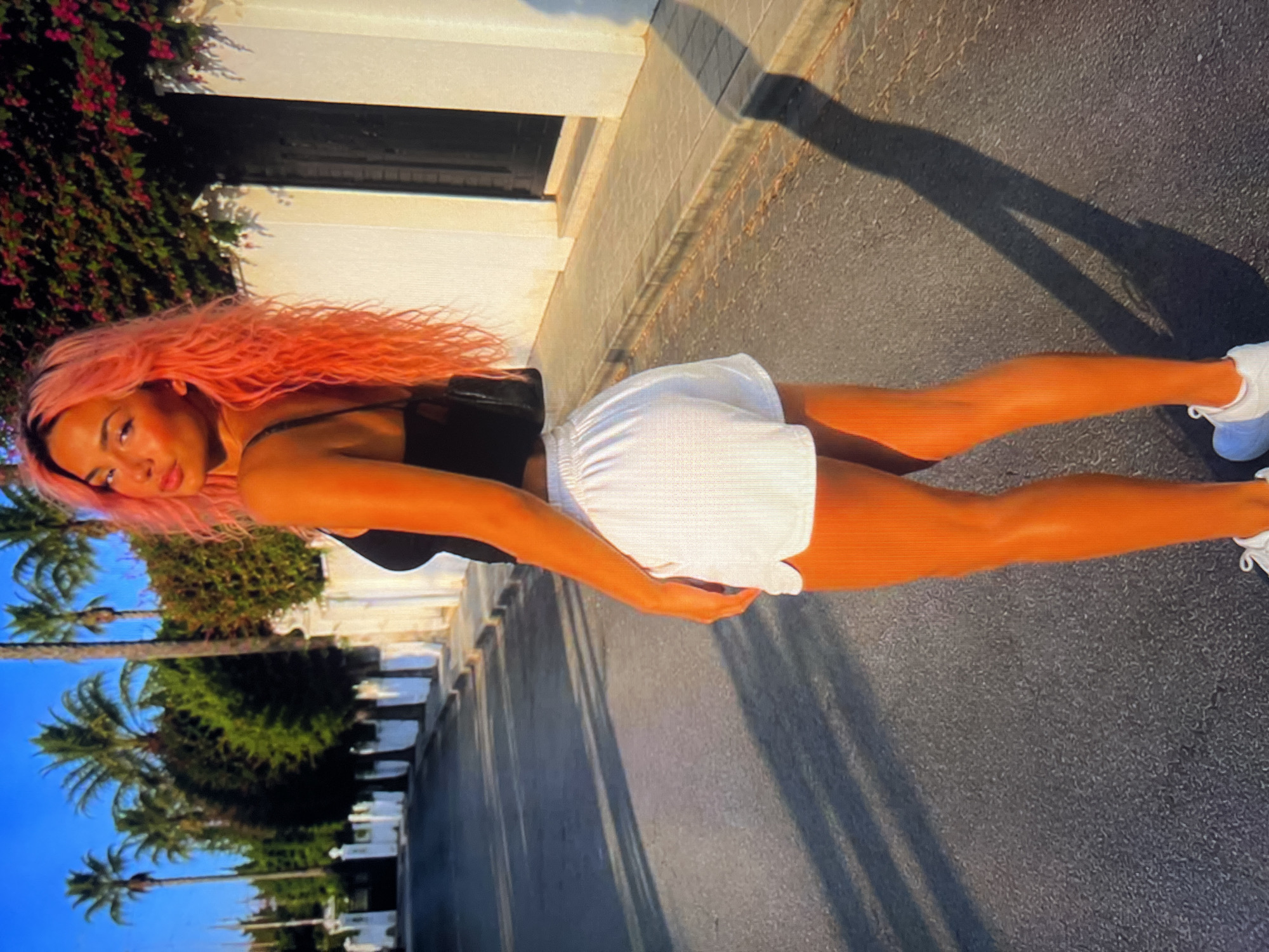 A young woman with vibrant pink hair stands on a paved street, facing away from the camera and looking over her right shoulder. She is wearing a black tank top and white athletic shorts. Her right leg is forward, and her left leg is bent at the knee, with her left foot slightly raised. She is wearing white athletic shoes. A black purse with a quilted pattern hangs from her left shoulder. The street is lined with a white fence and palm trees. The sky is a clear blue, and the sun is casting long shadows across the street. The overall impression is of a warm, sunny day in a tropical or Mediterranean-style location.