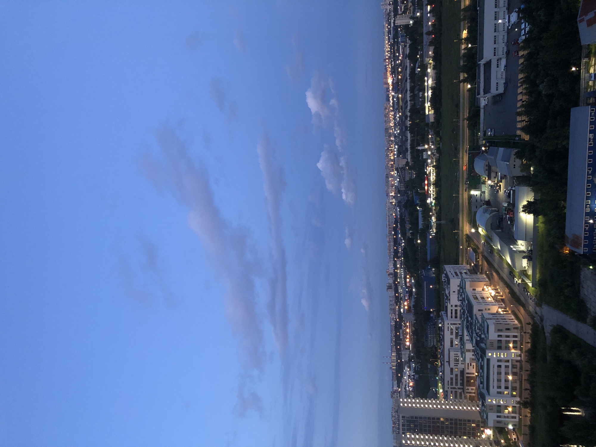 This is a high-angle, wide shot of a city skyline at dusk or dawn. The sky is a deep blue with scattered, wispy clouds. In the foreground, there are several modern, multi-story buildings with white facades and illuminated windows. The buildings are arranged in a cluster, with some appearing to be residential and others commercial. A road runs between some of the buildings, with streetlights illuminating the area.
Further in the distance, the city stretches out, a dense collection of buildings with countless lights twinkling, indicating a populated urban area. There are some taller structures, like smokestacks or industrial chimneys, visible on the horizon. The overall environment appears to be a developing urban area with a mix of residential and industrial zones. No people are discernible in the image. The lighting suggests it is either early morning or late evening, with artificial lights from the city beginning to dominate as natural light fades.