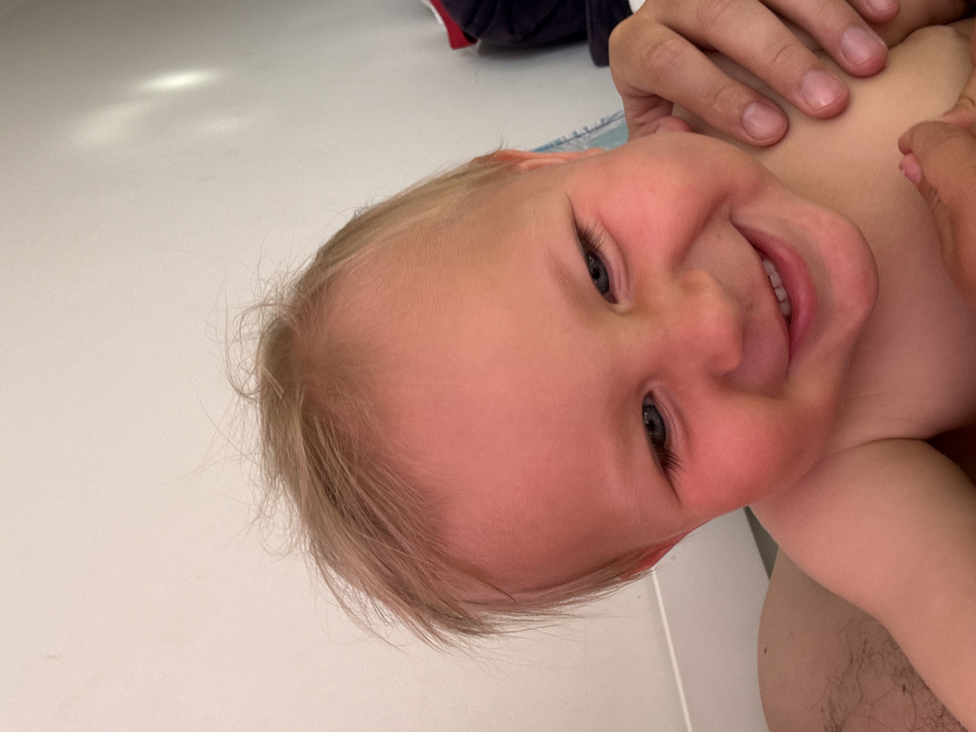 The image is a close-up, upward-facing shot of a young child. The child, with fair hair and rosy cheeks, is looking towards the camera with a wide smile, showing a few teeth. Their right eye is visible, appearing dark blue or gray. A person's hand is gently resting on the child's head and shoulder. The background is out of focus, appearing to be a light-colored, smooth surface, possibly a floor or wall, with some indistinct light reflections. The overall mood is playful and affectionate. There is no visible text. The location context is Sochi, Russia.