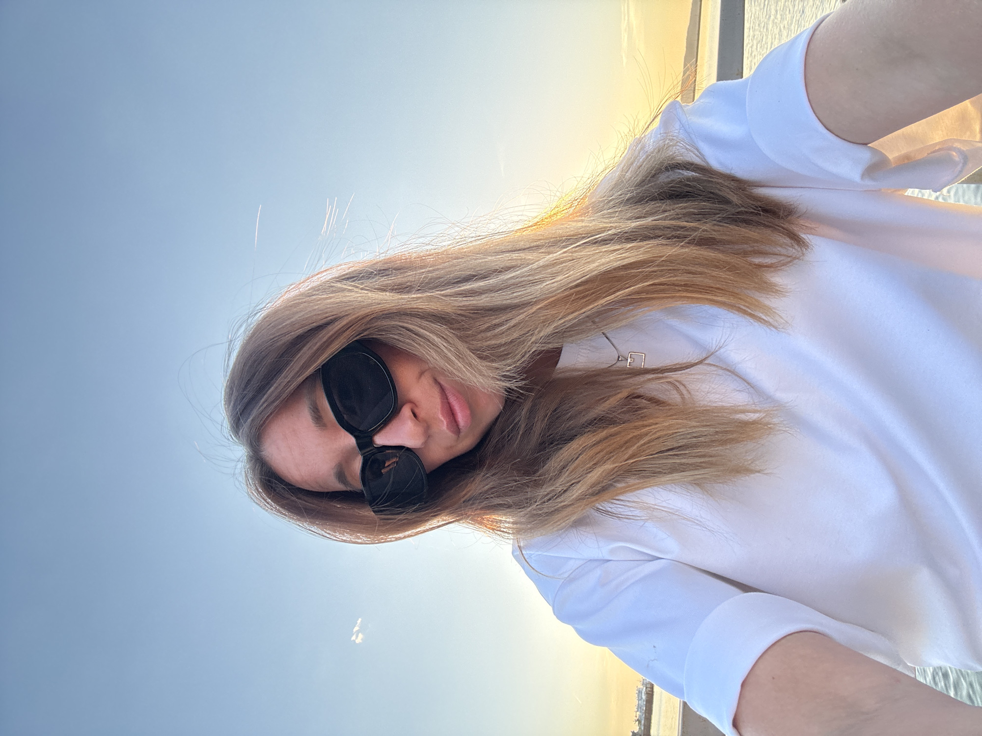 A woman is taking a selfie outdoors, wearing sunglasses and a white shirt. She has long, light brown hair. The background features a blue sky transitioning to a golden hue near the horizon, suggesting it is sunrise or sunset. In the distance, what appears to be a body of water is visible. A city with buildings can be seen in the far distance. While the image's origin is stated as Chaikovsky, Russia, this cannot be verified from the image alone.