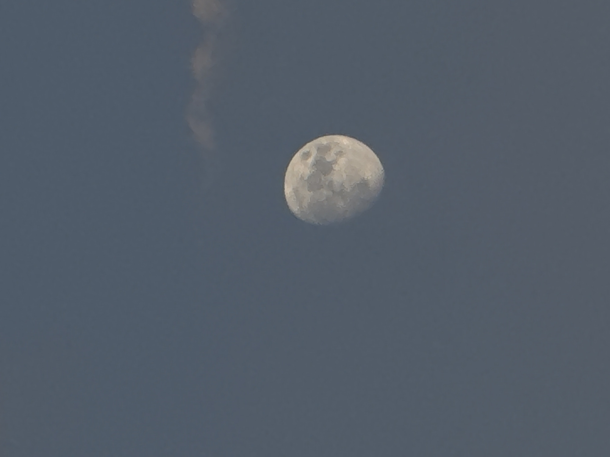 A close-up, vertical, eye-level shot features a gibbous moon against a dark, twilight sky. The moon, a pale white orb, is positioned in the lower right quadrant of the frame, displaying visible craters and textured surface detail. A wispy, faint cloud formation is present in the upper right portion of the image, partially obscuring the sky. The overall color palette is dominated by deep blues and grays, characteristic of a dusk or early evening setting. The location is not discernible from the image itself.