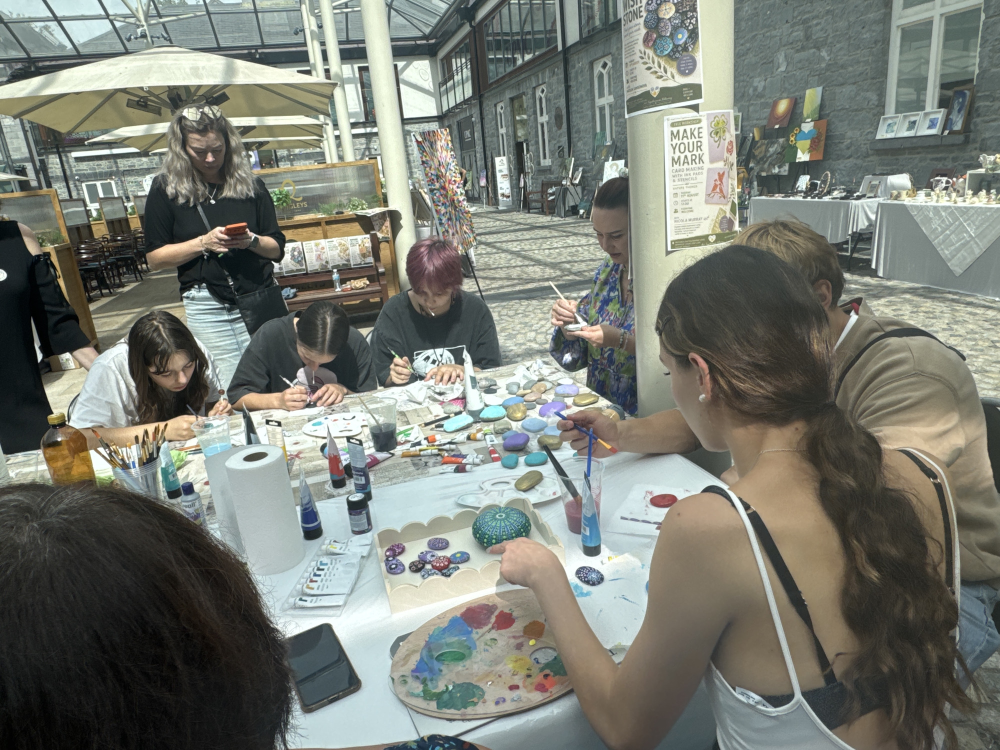 The image depicts an outdoor arts and crafts workshop, likely at the Kilkenny Design Centre in Kilkenny, Ireland. The setting features a covered outdoor area with a glass roof, providing natural light. Several individuals, predominantly young people, are gathered around tables, engaged in painting small stones.
On the table, a variety of art supplies are visible, including tubes of paint, paint palettes, brushes in various containers, water cups, and rolls of paper towels. Numerous painted stones, some decorated with intricate patterns and designs, are scattered across the table and arranged in a wooden tray. One person in the foreground, with long brown hair tied in a ponytail, is actively painting a green stone. Other participants are also shown actively painting or preparing their stones.
In the background, there are displays of artwork and crafts, suggesting this is part of a larger exhibition or market. A woman in a black top and a denim skirt is visible in the upper left, looking at her phone. The architecture of the building, characterized by stone walls and large windows, is consistent with the historic buildings found in Kilkenny. The overall atmosphere suggests a creative and engaging community event.