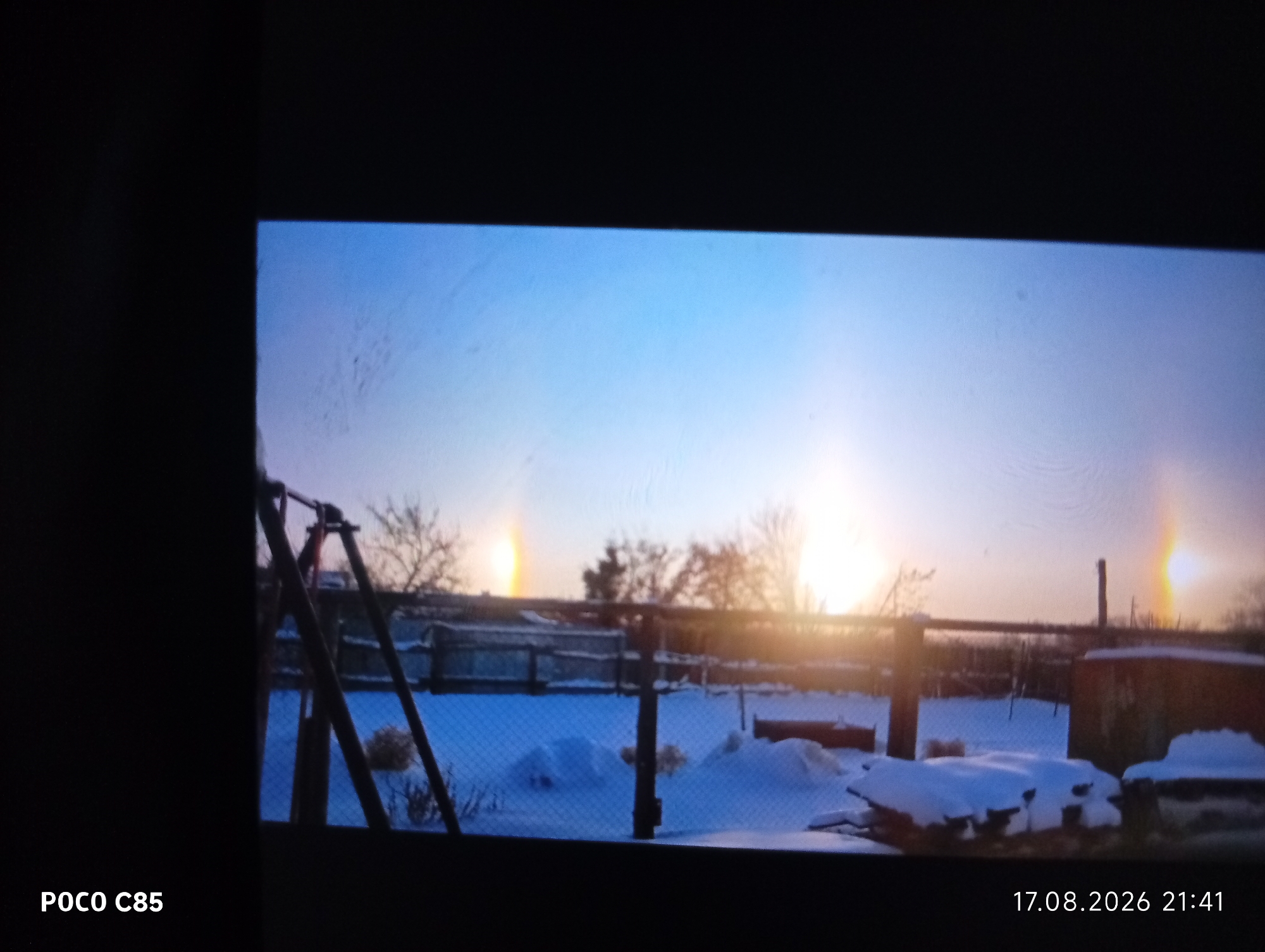 The image shows a wintry scene in a rural area, likely a backyard, with snow covering the ground and piles of snow visible. There is a wire fence in the foreground and trees in the background. The sky is a gradient of blue and white, with bright spots of light, possibly sun dogs, visible on either side of a central bright area, suggesting a sunset or sunrise. A metal swing set structure is partially visible on the left side of the image. The timestamp in the lower right corner reads "17.08.2026 21:41," and "POCO C85" is in the lower left corner, indicating the device used to take the photo. The scene appears to be set in Stepanovka, Kazakhstan, as indicated by the prompt.