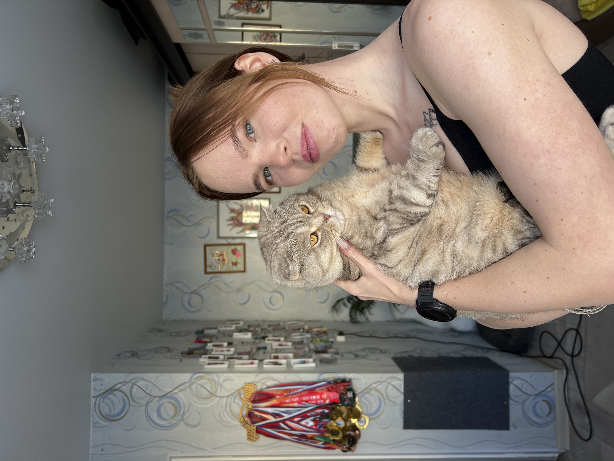 A woman is holding a tan and grey tabby cat. She is wearing a black tank top and a black sports watch. In the background is a chandelier on a grey wall, a mirror with various small framed pictures, a wall with a patterned wallpaper, a table with small boxes stacked on top and a set of medals attached to the wall. The location of the photograph is Chaikovsky, Russia.