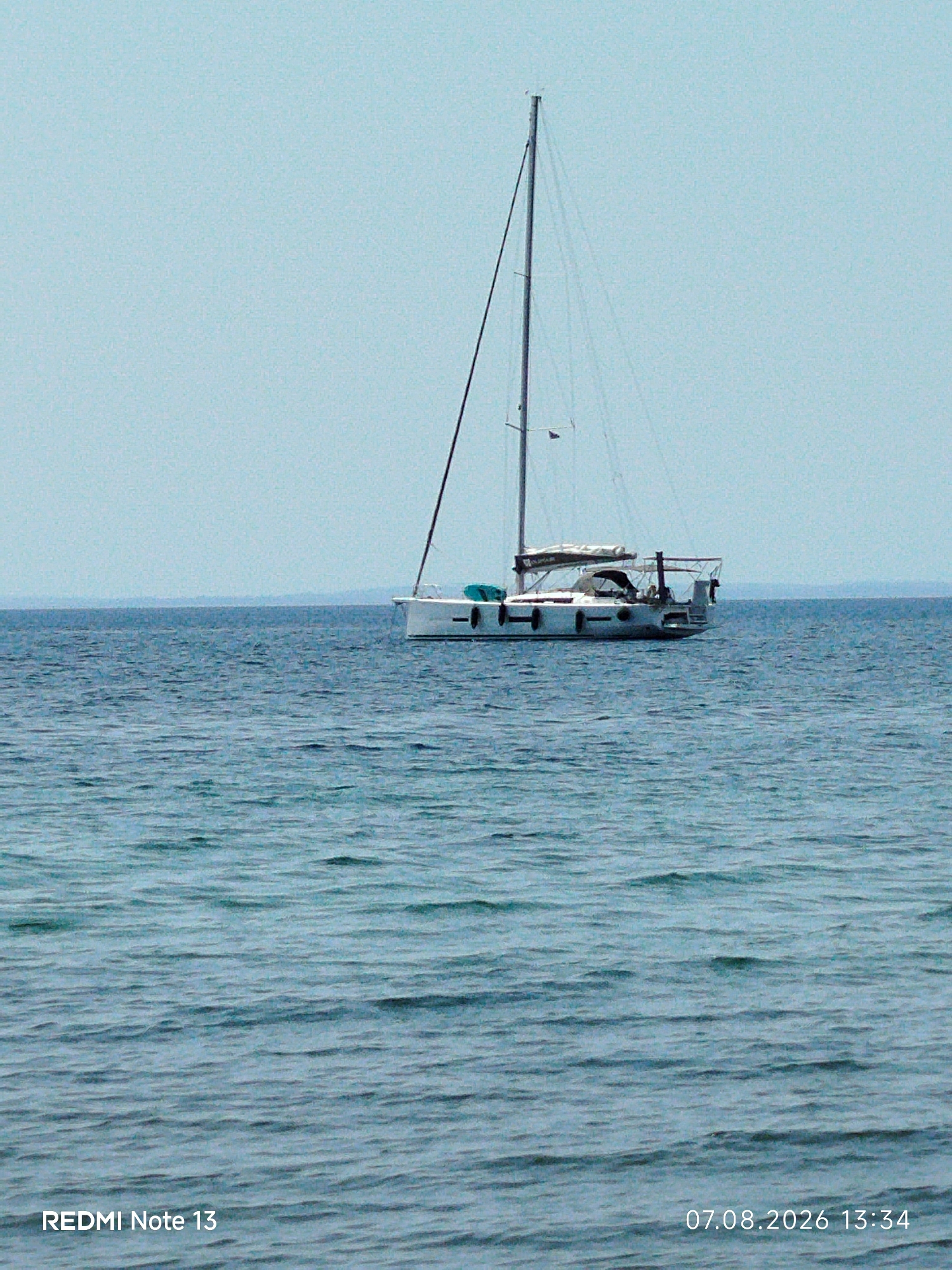 A single sailboat is pictured at sea under a clear sky. The sailboat is white with a dark mast and rigging. It is positioned on the right side of the image, facing left. The water is a deep blue with small ripples. In the distance, a faint line of land is visible on the horizon. The bottom left corner of the image displays "REDMI Note 13" and the bottom right corner shows "07.08.2026 13:34".