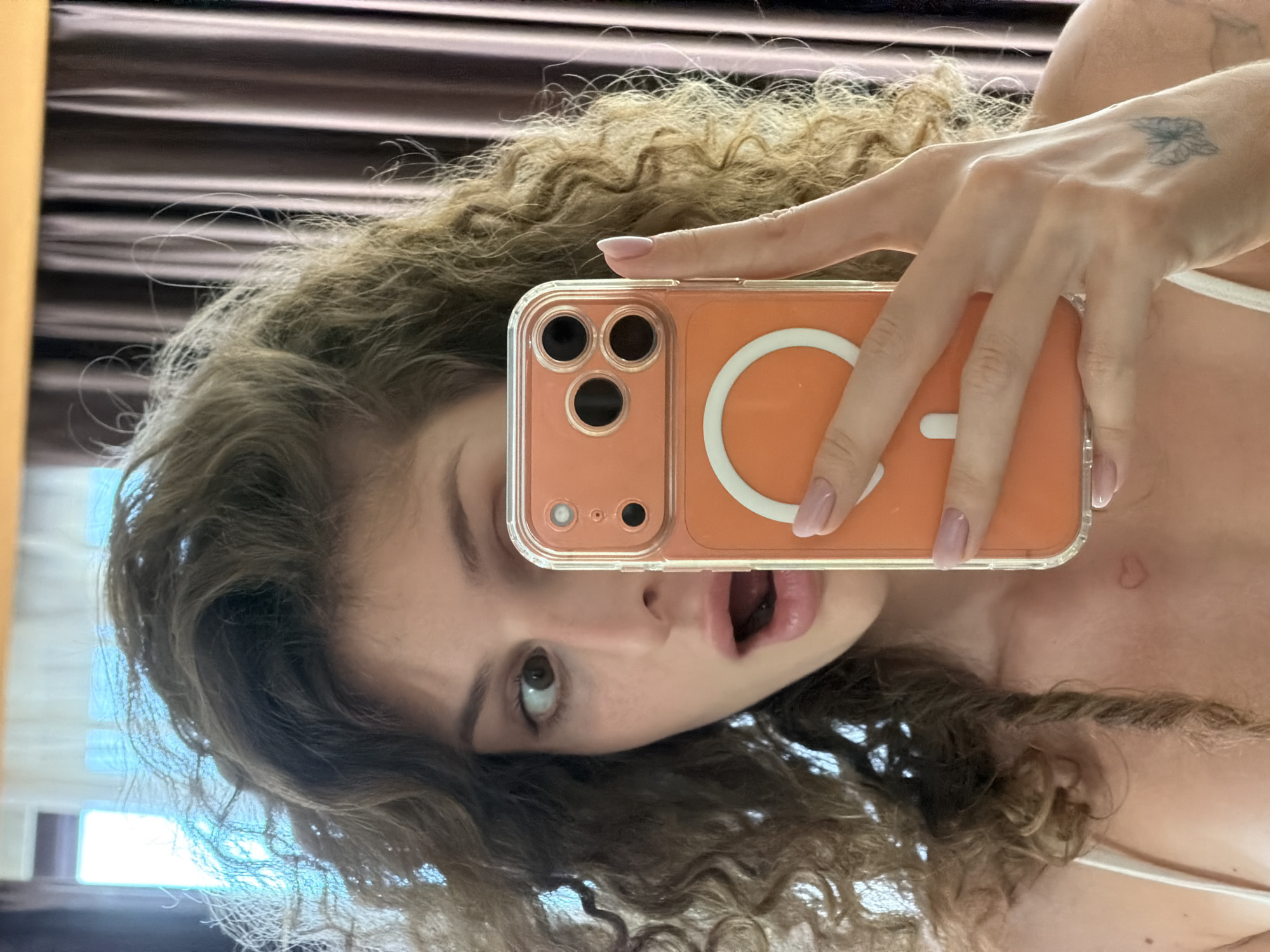 A fair-skinned person with curly brown hair is holding an orange iPhone in front of their face, taking a selfie. The person has light-colored fingernails. The phone has a clear case with a white circle on the back. Behind the person is a blurred background with a curtain, and a window with diffused sunlight. The location is not explicitly identifiable as North Kuta, Indonesia from the visual information provided.