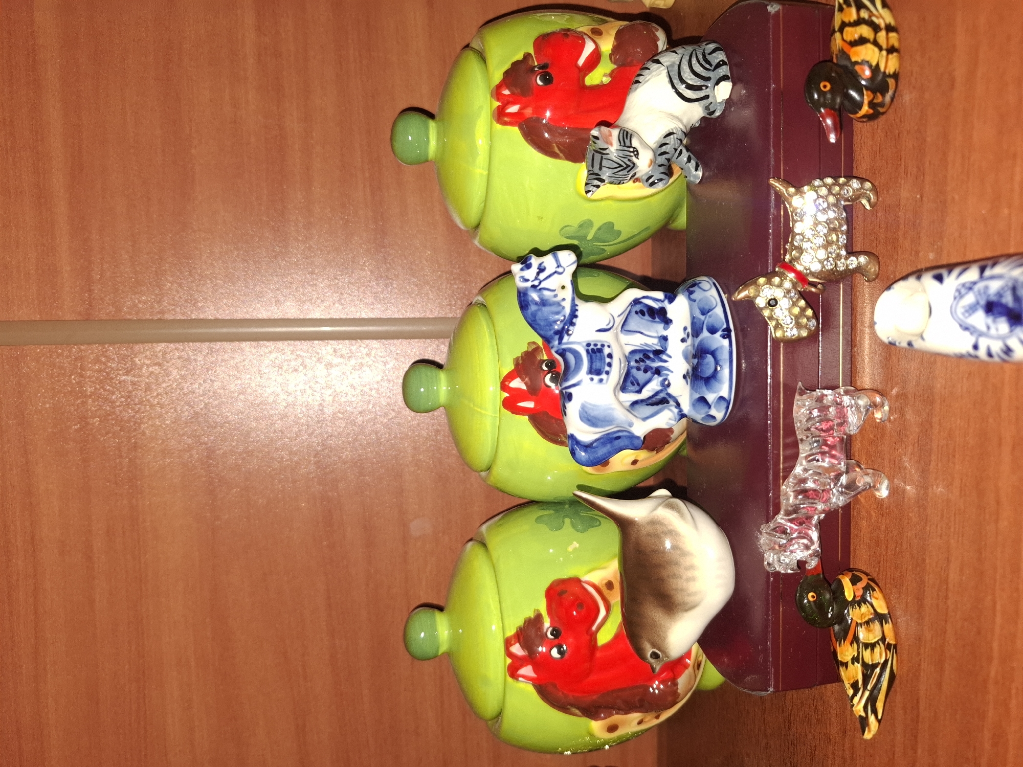 The image displays a collection of ceramic figurines and decorative objects arranged on a wooden surface, likely a shelf or table. In the background, a wooden wall with horizontal grain is visible. The arrangement includes three large, lime-green ceramic jars, each adorned with a painted red horse figure. In front of these jars are smaller figurines: a brown and white bird, a blue and white ceramic horse figurine, and a grey and white striped cat figurine. In the foreground, several smaller decorative items are placed, including two duck figurines, a clear crystal figurine of a dog, and a small jeweled figurine of a dog wearing a red collar. A small, white ceramic object with blue patterns is also visible in the foreground. The lighting suggests an indoor setting. The image does not provide any information about the city of Almaty, Kazakhstan.