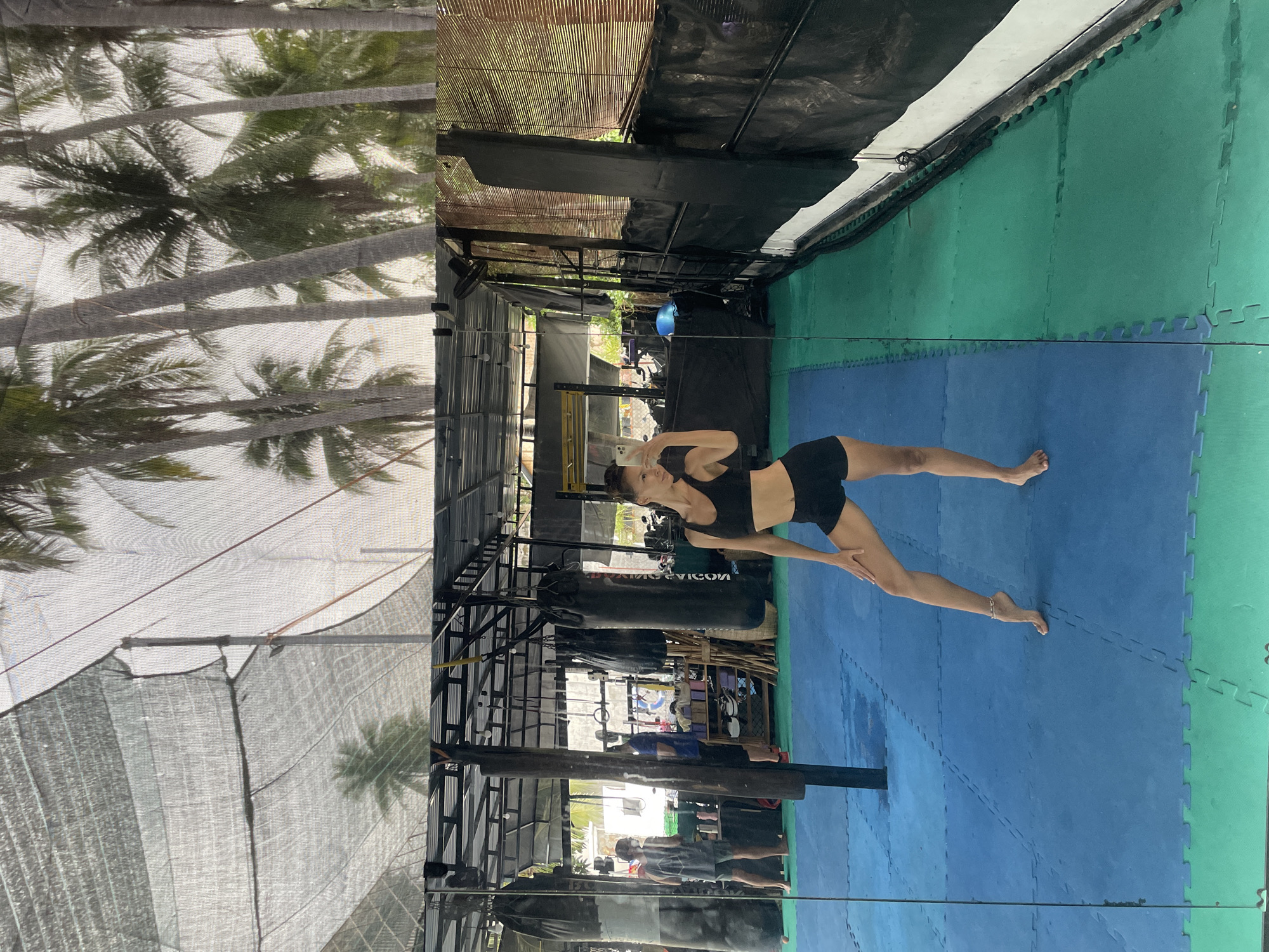 The image, correctly oriented, depicts a slender woman in a semi-outdoor gym or training facility, likely located in Phan Thiet Ward, Vietnam. She is barefoot, dressed in a black sports bra and black shorts, striking an athletic or yoga-like pose with her left leg extended backward and her left arm raised. A reflection, possibly from a large mirror or glossy surface, shows her holding a smartphone up, suggesting she is capturing her pose.
The floor is covered with blue and green interlocking puzzle mats. The gym environment features various equipment, including multiple black punching bags, one prominently displaying the text "KOVICON". Metal scaffolding, pull-up bars, ropes, and a mounted fan are also visible. The roof structure is a combination of dark tarpaulin and natural bamboo slats.
Beyond the immediate training area, lush green palm trees are visible through a light-colored mesh or shade netting, indicating a tropical, open-air setting. The bright natural light suggests it is daytime. Other individuals are faintly visible in the background, some as reflections, engaged in various gym activities. The scene conveys a focused training session within a vibrant, outdoor-integrated environment.