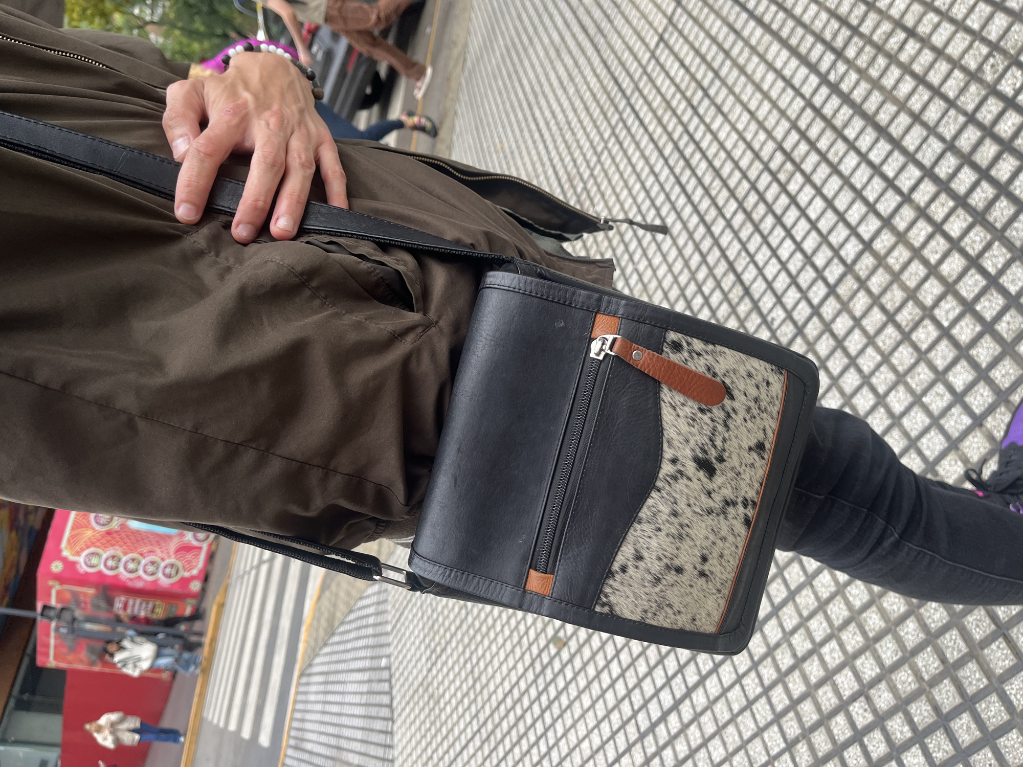 The image presents a first-person perspective view looking downwards, likely in an urban outdoor setting in Buenos Aires, Argentina. The primary subject is a person wearing a dark olive or brown jacket, with a black cross-body strap extending across their chest. Their left hand, adorned with a bracelet of white and dark beads, rests casually on the jacket near the strap.
Attached to the strap is a distinct small cross-body bag. The bag is made of black leather, featuring a prominent section of mottled white and black cowhide-like material. It has brown leather accents, including a small pull tab for a silver zipper located on the front of the bag.
The ground beneath is paved with light-colored square or rectangular tiles, and strong sunlight casts a sharp, repetitive grid-like shadow pattern across the surface, indicating a clear, sunny day.
In the blurred background, other elements of an urban environment are visible. These include faint, out-of-focus figures of other pedestrians, one wearing black jeans and partially visible purple shoes near the bag, and another in a light jacket and blue jeans further back. A blurry red structure, possibly a kiosk or building facade with indistinct graphics, can also be seen in the distance. No legible text is visible in the image.