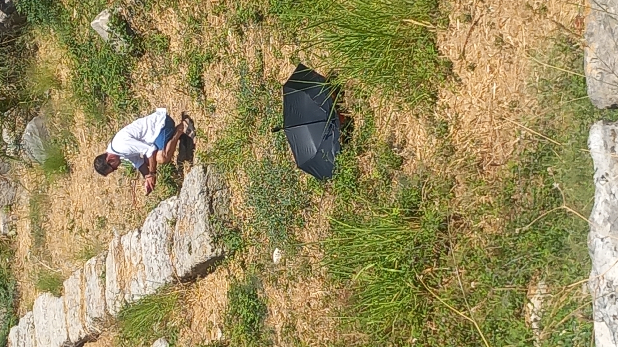 A person wearing sunglasses, a white short-sleeved shirt, and blue shorts is crouching on a grassy slope in the city of Kos, Greece. The person is looking at a smartphone held in their hands. To the right of the person, a black umbrella is resting on the ground. The background consists of a stone pathway and dry vegetation.