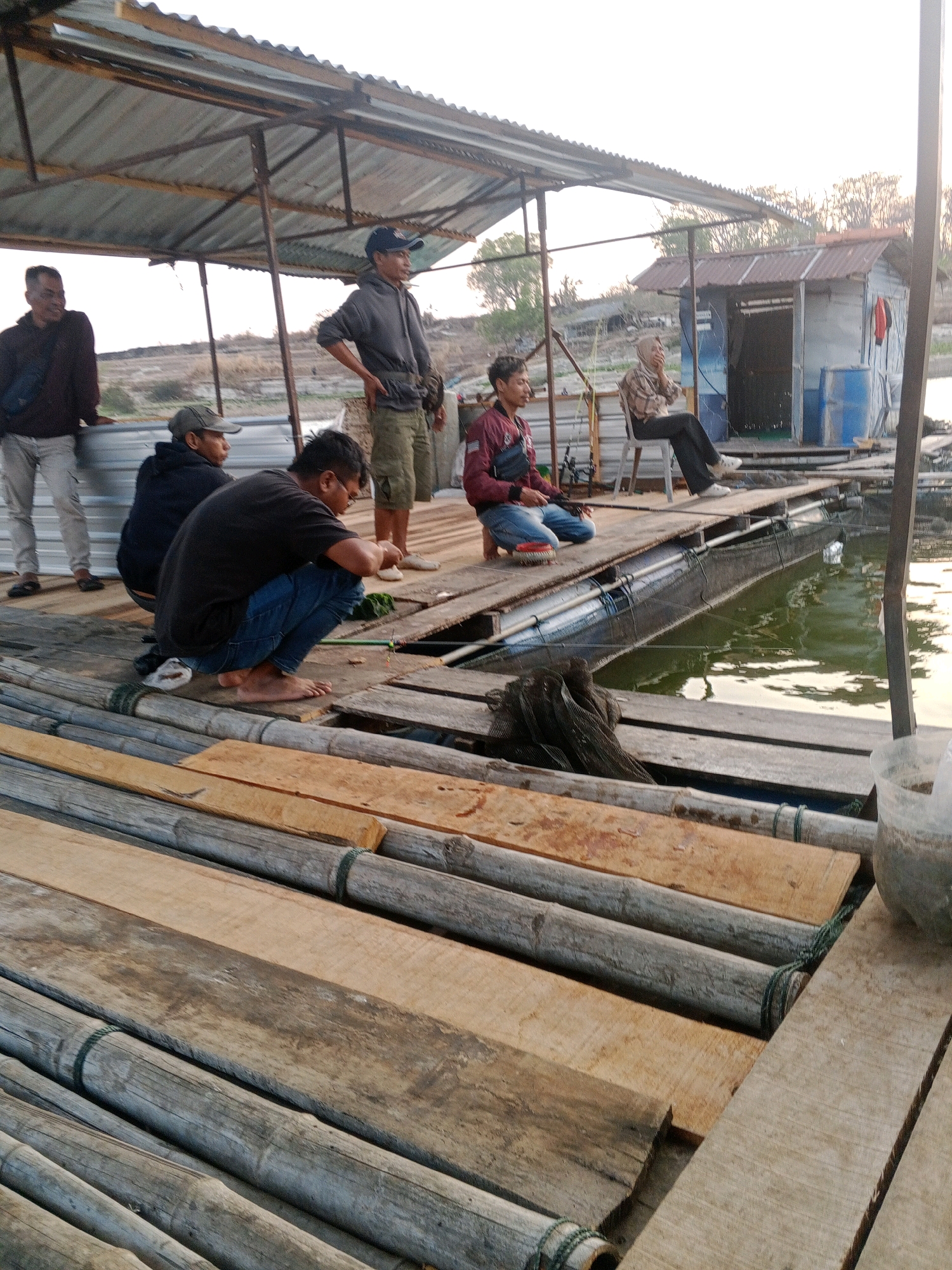In Sendang, Indonesia, a group of men are seen on a floating wooden platform by a body of water. The platform is constructed from logs and planks, with a makeshift roof overhead, possibly for shade. One man, wearing a dark hoodie and a baseball cap, stands and observes. Another man, in a grey hoodie and also wearing a baseball cap, is seated on the platform. A third man, in a black t-shirt and blue jeans, is crouched down, appearing to be fishing. Further back, another man in a red jacket is also engaged in fishing. In the background, a small shack-like structure with a blue barrel and orange life preserver is visible, along with trees and a hazy sky. The overall scene depicts a fishing or recreational gathering on a water body.