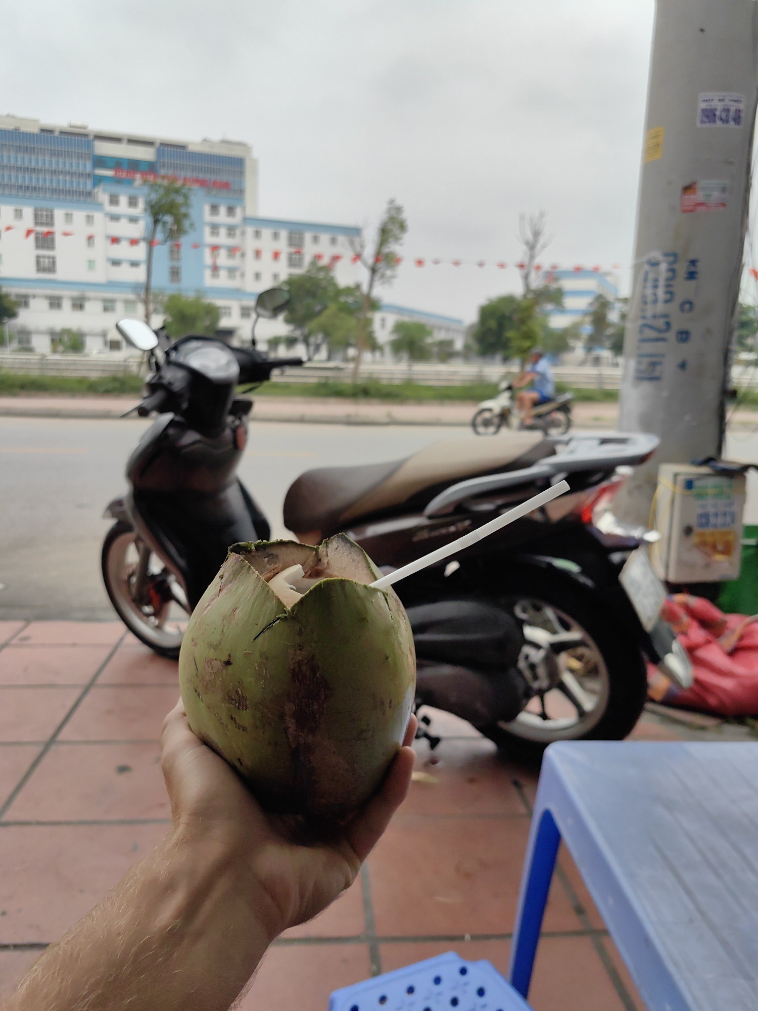 A person holds a green coconut with a white straw in front of a black scooter on a red-tiled sidewalk in Cao Xanh Ward, Vietnam. In the background, a multi-story building with white and blue accents is visible under an overcast sky. A utility pole stands to the right, marked with writing and stickers. Another scooter is visible on the road behind the first. A blue plastic table is partially visible in the foreground.