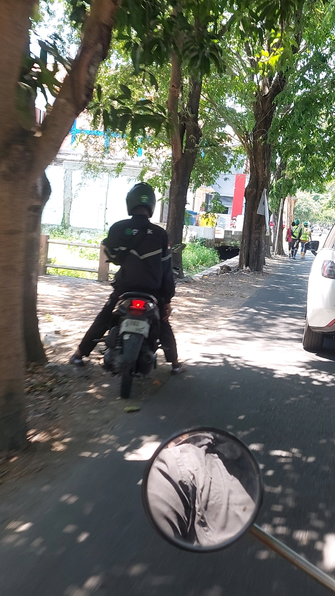 In Siwalan Panji, Indonesia, a person wearing a dark uniform and a green helmet sits on a black motorcycle parked by the side of a paved road. The motorcycle's license plate displays the number "6102." Behind the parked motorcycle, two individuals in high-visibility vests and helmets stand on the roadside, appearing to be workers. The environment is characterized by the presence of mature trees lining the road, casting shadows on the asphalt. A white car is visible on the right side of the road, and in the background, a building with blue and white accents can be seen. The scene suggests a bright, daytime setting with filtered sunlight.