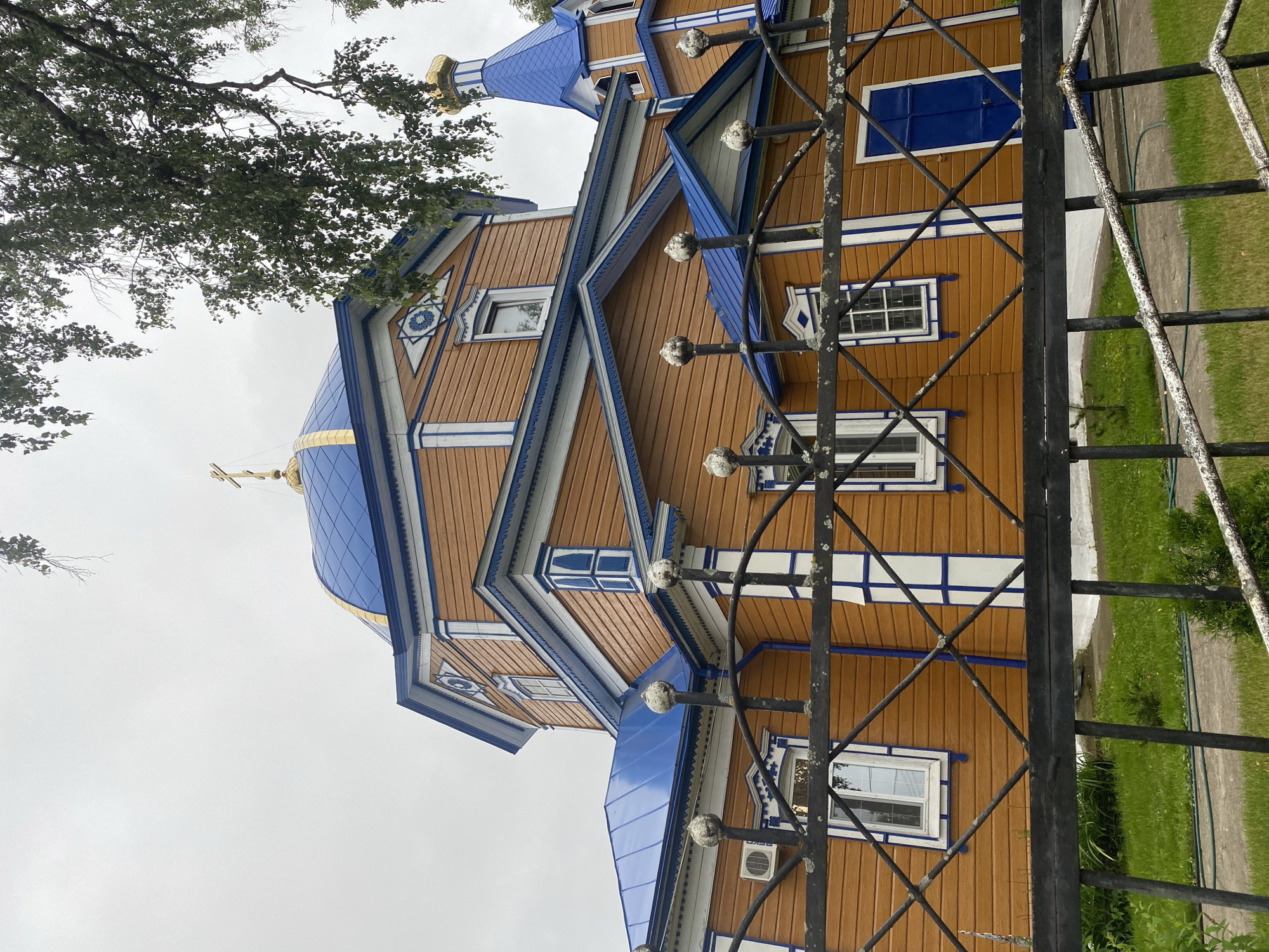 The image shows the exterior of an Eastern Orthodox church in Zelenodol'sk, Russia, on an overcast day. The church has a wooden exterior with orange siding and blue roofs, accented with white trim and blue decorative elements. It features a large blue dome topped with a golden cross, and a smaller bell tower with a golden dome. A black metal fence with ornamental posts is in the foreground, obscuring a portion of the church's lower level. Beyond the fence, a green lawn and a paved pathway are visible. The scene is devoid of any people or animals.