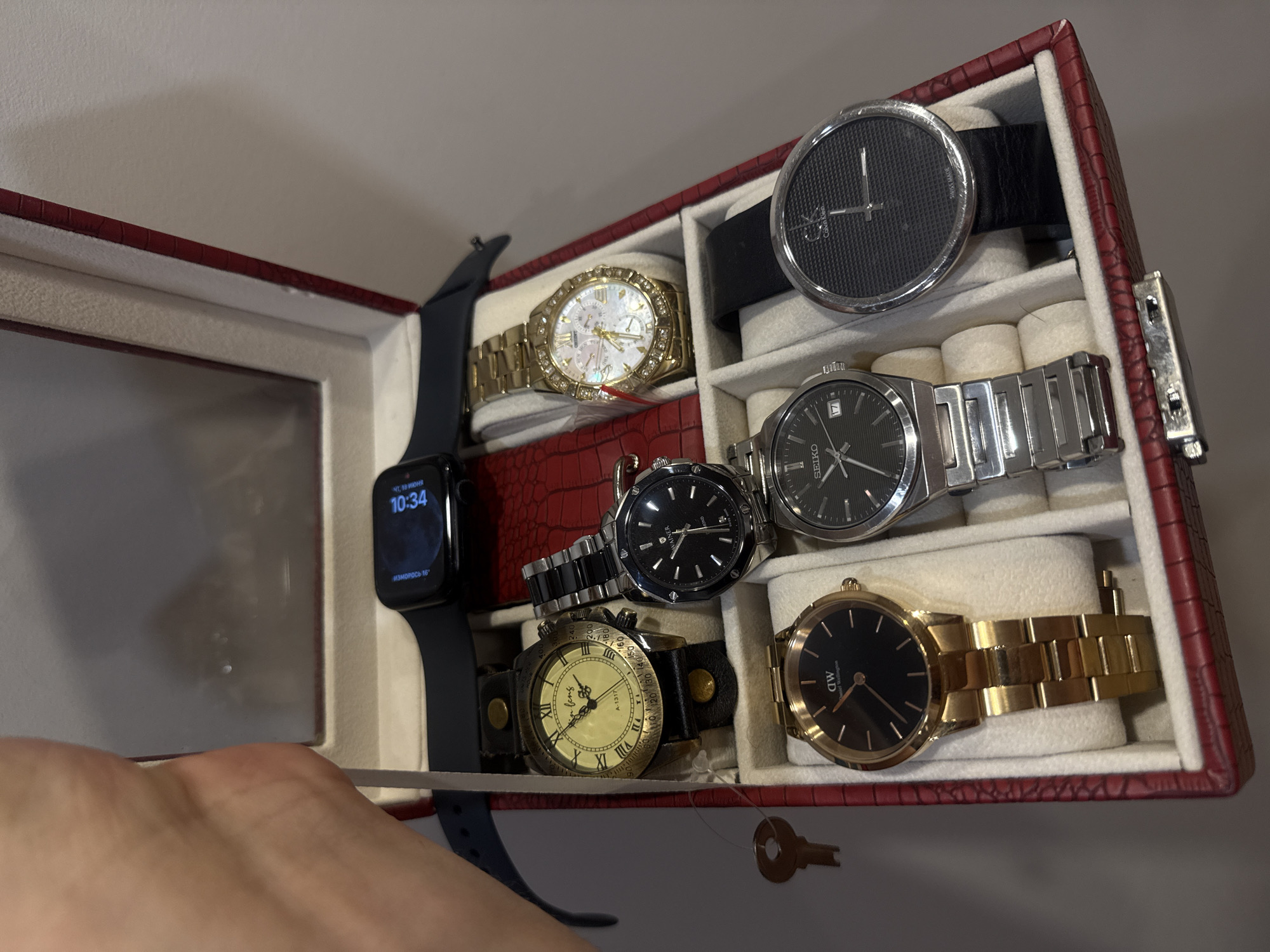 The image shows a watch organizer box, which appears to be made of red crocodile-embossed leather, containing several watches. From left to right, top to bottom, the watches are:
1. An Apple Watch with a black band, displaying the time "10:34" and a moon phase icon.
2. A vintage-style watch with a cream-colored dial, Roman numerals, and a dark leather strap. The dial has Chinese characters and "A-1317" printed on it.
3. A gold-tone watch with a mother-of-pearl dial, multiple sub-dials, and a bezel decorated with small stones.
4. A black-faced watch with a silver-tone metal band and the brand name "WAINER" visible on the dial.
5. A gold-tone watch with a black dial and a "daniel wellington" logo.
6. A silver-tone watch with a black dial, the brand "SEIKO" and a date window showing "17".
7. A watch with a black leather strap and a round, dark gray dial with a textured pattern and the "Calvin Klein" logo.
A small, brass-colored key is visible to the left of the organizer box. The organizer is placed on a neutral-colored surface. The background is out of focus but suggests an indoor setting with a wall. There is a hand in the upper left corner of the image, holding the edge of the organizer. There is no information in the image that suggests the location is in Vladivostok, Russia.