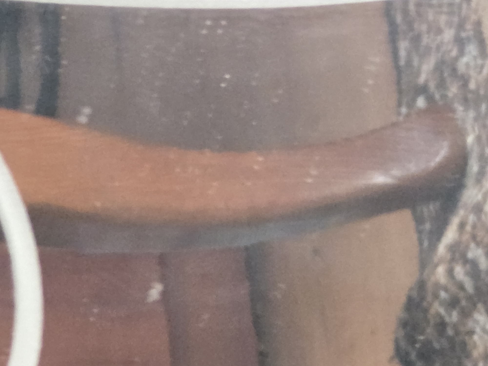 A close-up shot focuses on the lower leg and foot of a person, possibly a child, standing on a sandy or gravelly surface. The leg is light brown in color and appears smooth. A white object, possibly a railing or bar, is visible across the top of the image. The background is a muted reddish-brown with darker horizontal lines, suggesting a textured wall or surface. The lighting is soft, casting gentle highlights on the skin. The image is pixelated, making precise identification of details challenging. The scene is set in an outdoor environment, possibly a park or a recreational area within the city of Unknown, Unknown.