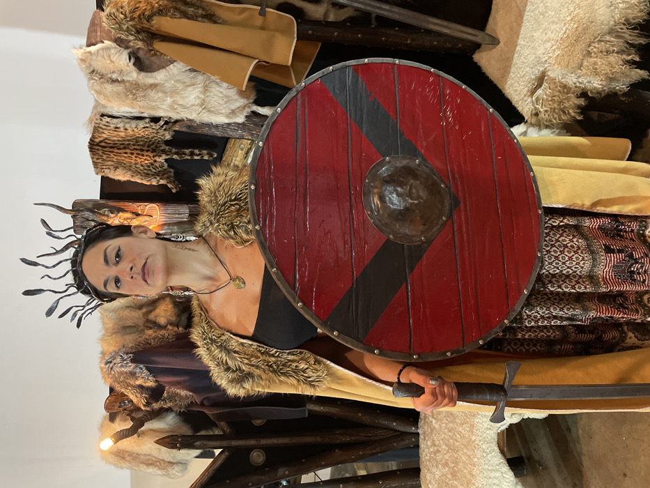 A woman dressed as a Viking holds a sword and a red and black shield. She wears a fur-trimmed outfit, a black top, and a unique crown-like headpiece. Various furs are draped behind her, along with a tan garment. The scene appears to be part of a staged display or a historical reenactment, lit to highlight the textures and colors of the Viking attire and accessories.