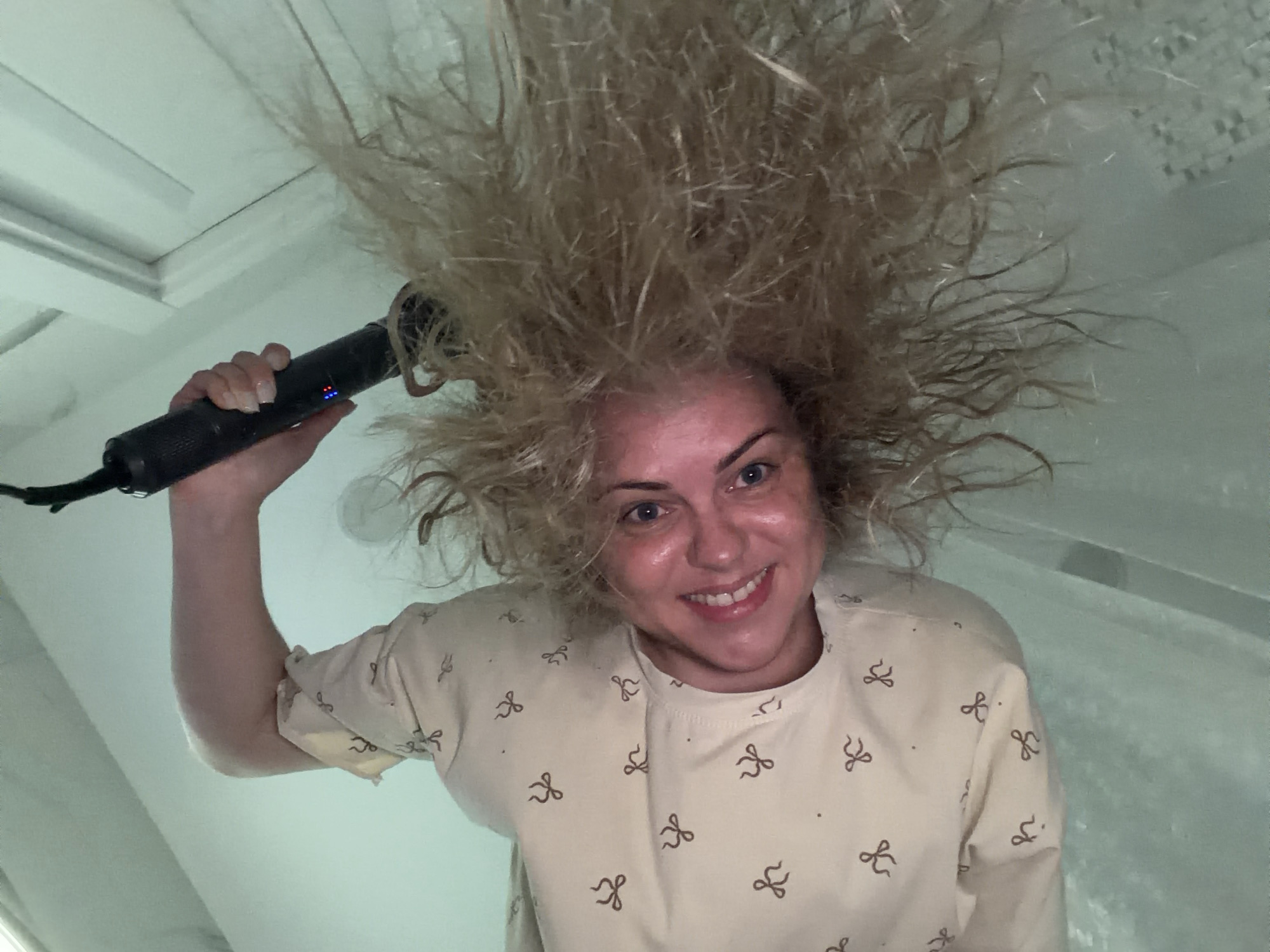 A woman with blonde, frizzy hair is smiling at the camera while holding a black hair styling tool above her head. The woman has fair skin and blue eyes, and is wearing a beige top with a bow pattern. The background appears to be a white interior space, possibly a bathroom, with elements like a ceiling with molding visible. There is no information in the image to identify Votkinsk, Russia, or any other specific city.