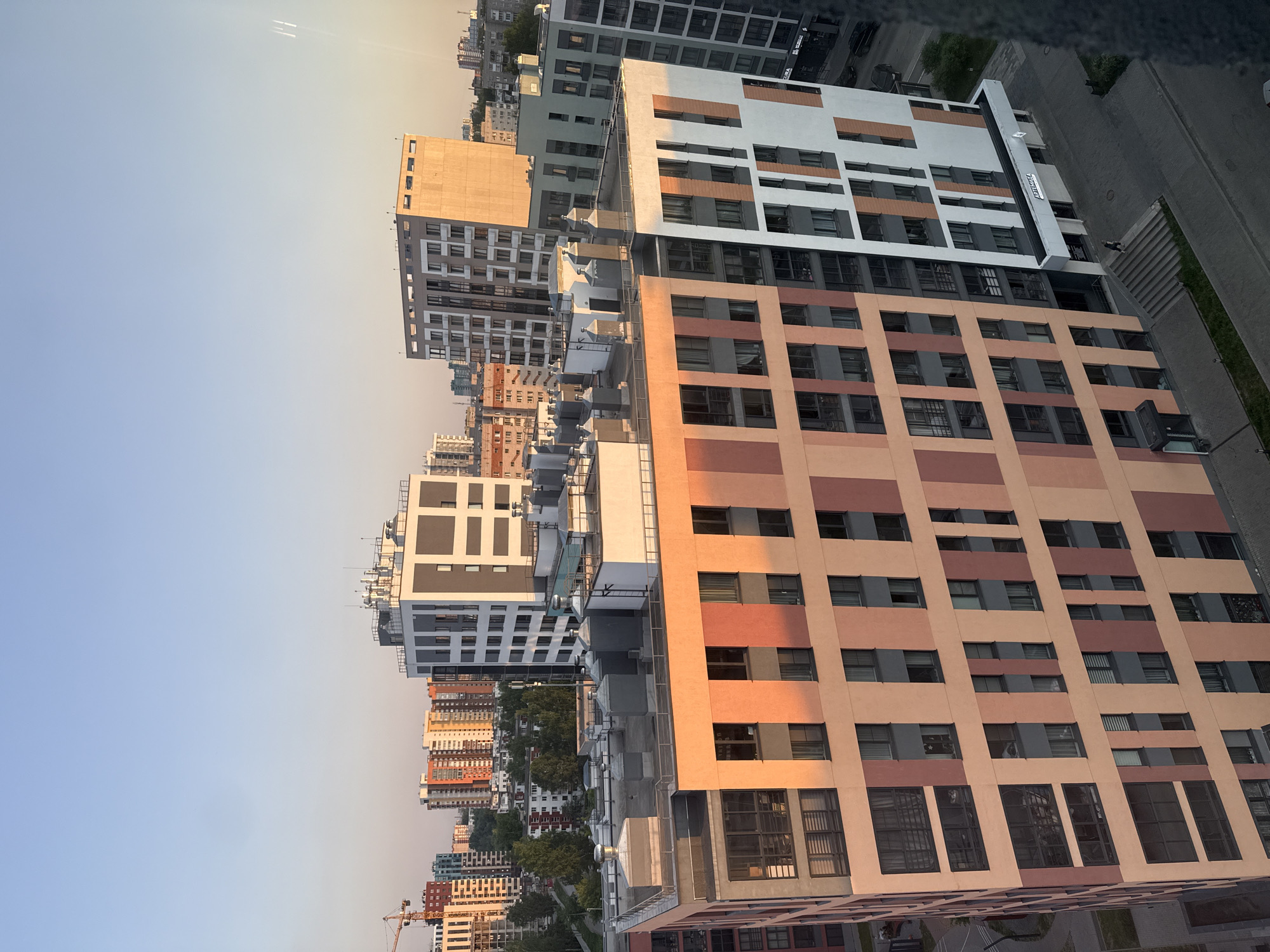 The image shows a high-angle view of a cityscape, likely taken from a tall building, with a clear sky in the upper left corner. Several buildings of various heights and architectural styles dominate the view. Many of the buildings feature a grid-like pattern with windows arranged in rows and columns. Some buildings are painted in light shades of white, beige, and peach. A building with a sign that reads "Paradigma" is visible, which suggests that this is Izhevsk, Russia. In the lower left corner, a construction crane is also seen, suggesting ongoing urban development. The image exhibits a warm, golden-hour lighting that casts long shadows.