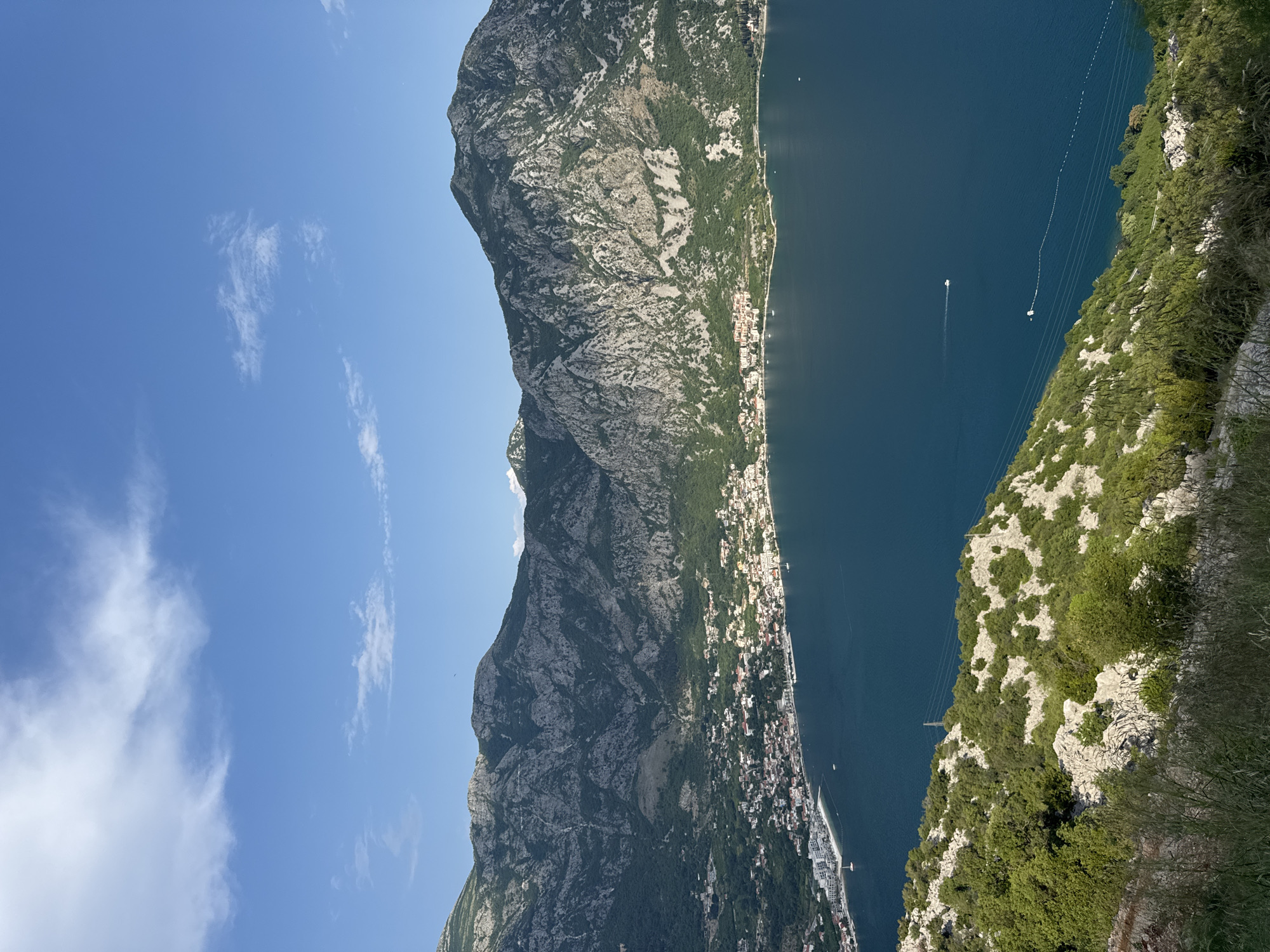 An aerial, wide-angle shot captures the Bay of Kotor in Strp, Montenegro, under a clear blue sky with scattered white clouds. The bay's deep blue water is in the foreground and mid-ground, with several small white boats visible on its surface. On the left side of the bay, a coastal town with white and orange buildings lines the shore, backed by a steep, green, and rocky mountainside. The mountains themselves are rugged and grey, with patches of green vegetation clinging to their slopes. The foreground of the image shows a rocky, green hillside with sparse vegetation. The lighting suggests it is daytime, with sunlight casting shadows on the mountains.