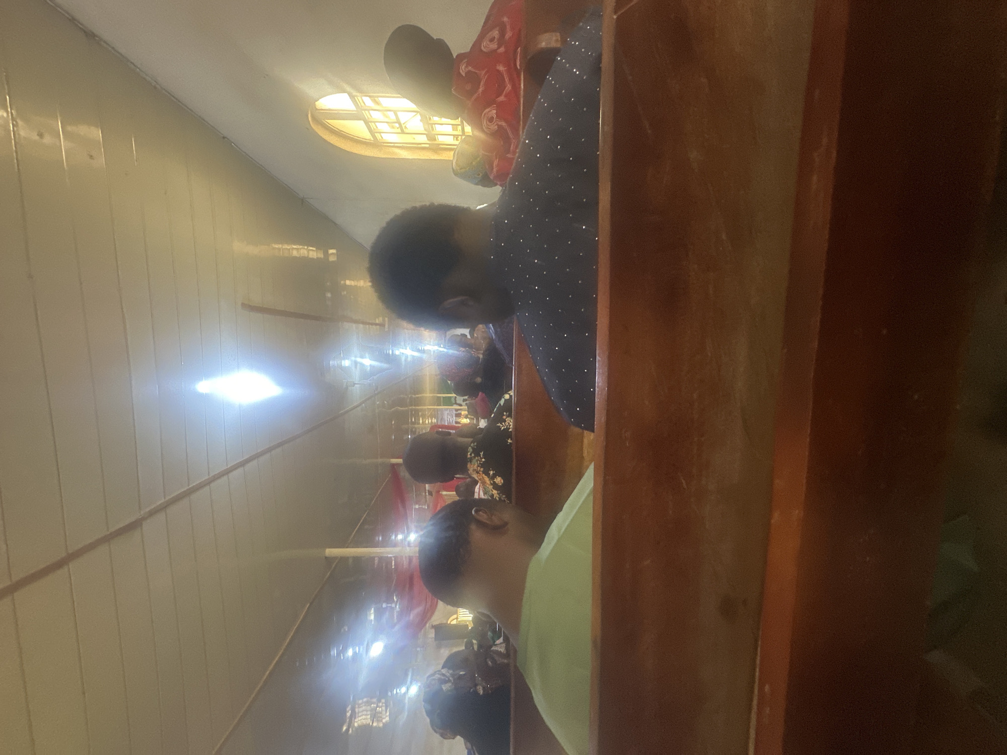 The image captures an indoor gathering in Jos, Nigeria, viewed from the perspective of a seated attendee. The foreground displays the backs of two wooden pews or benches. In the immediate foreground, a male individual wearing a light green t-shirt is seated on the left, while another male individual in a dark blue shirt with small, light-colored embellishments is seated on the right. Both individuals are viewed from behind their heads and shoulders. Further into the mid-ground, additional people are visible, also seated and facing forward. One individual on the far right is dressed in a red garment with white patterns. The ceiling, constructed from light-colored, rectangular panels, exhibits strong reflections from multiple overhead light sources, creating bright streaks. On the right-hand wall, a distinct arched window with a grid of panes allows bright external light to enter the space. Elements of the background include obscured architectural features and sections of draped red fabric.