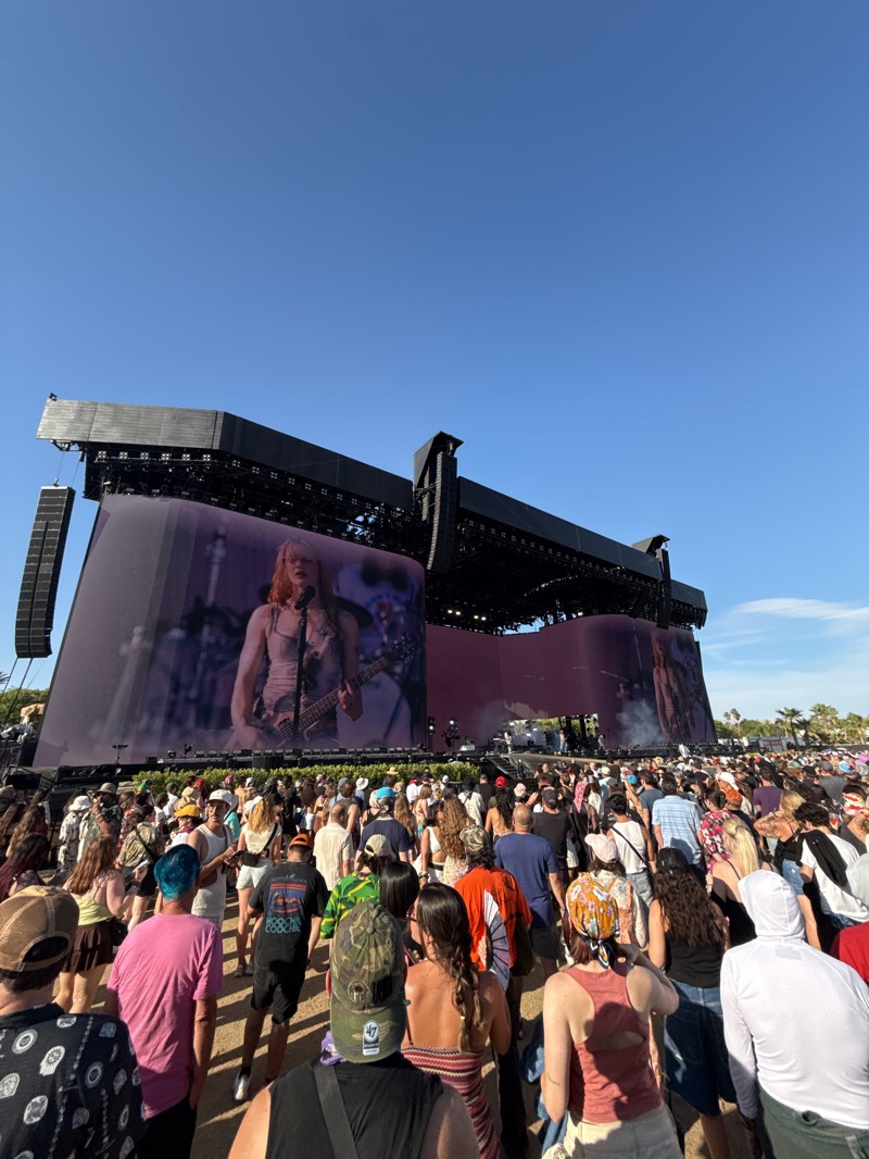 The image shows a large crowd of people gathered in front of a massive stage, likely at the Coachella Valley Music and Arts Festival in Indio, California, United States. The stage features large screens displaying a performer with long hair and a guitar. The sky is clear and blue, suggesting it is daytime. The crowd is dense, with people of various ages and styles of clothing. The scene appears to be a live concert or performance.