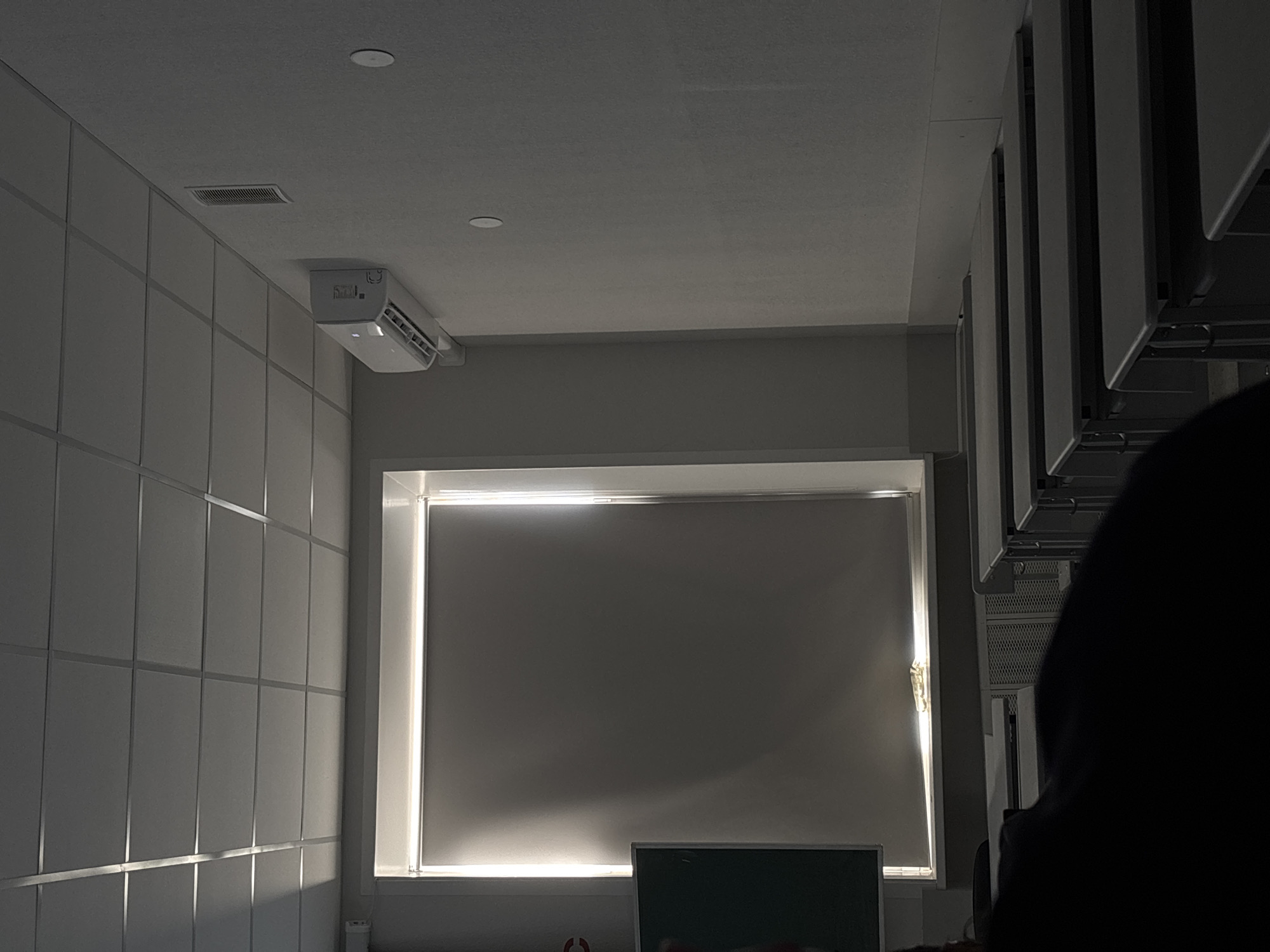 The photograph, taken indoors, presents a scene with a blend of ambient and artificial light. A window with a pull-down shade is the central focus, emitting a soft glow around its edges. Below the window, a portion of a dark object, possibly a screen or board, is visible. To the left of the window, a wall with vertically aligned panels catches the light, creating a pattern of shadows and highlights. An air conditioning unit is mounted near the ceiling in the upper-left corner. On the right, partially obscured, are vertical metal structures. A dark, ambiguous shape dominates the lower-right portion, obstructing a clearer view. The location is possibly in Perm, Russia, though there is no concrete evidence within the photograph to confirm it.