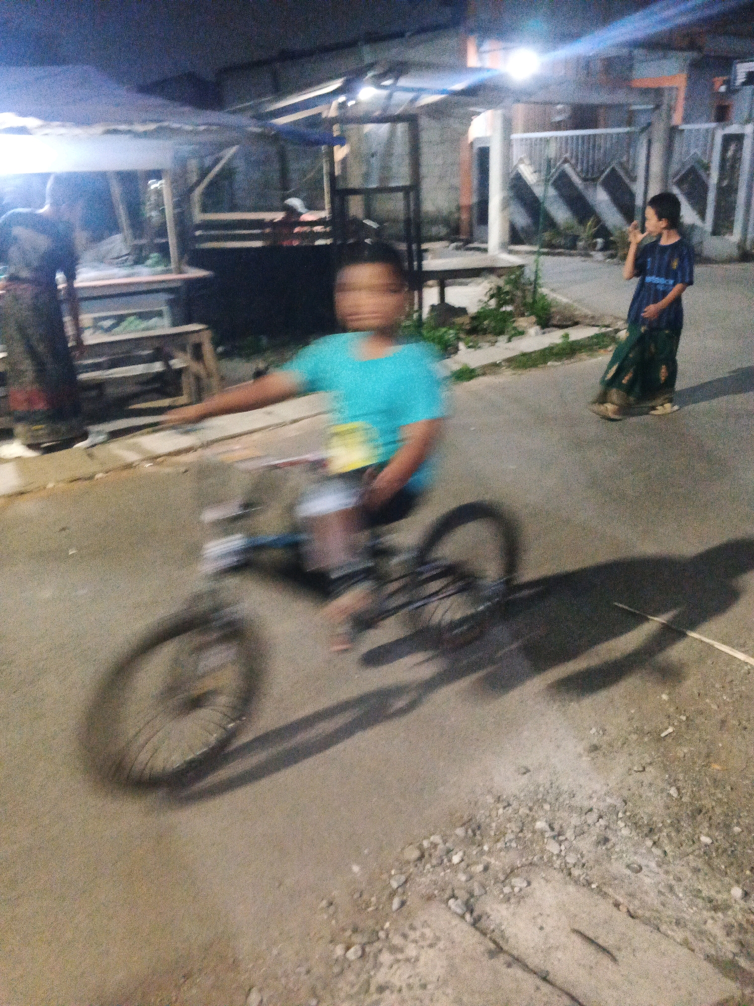 The image captures a motion-blurred scene at night on a paved street in Kadu, Indonesia. In the foreground, a young person is riding a bicycle, with their body and the bicycle appearing indistinct due to movement. The cyclist is wearing a turquoise t-shirt and dark shorts. To the right, a young person in a dark blue t-shirt and a patterned green skirt stands on the side of the road, looking to their right. Further in the background, another person in dark clothing is partially visible standing next to a wooden structure with a corrugated roof. The environment appears to be a semi-urban or rural setting, with the street flanked by simple structures and vegetation. Lighting appears to be from artificial sources, creating shadows and illuminating parts of the scene.