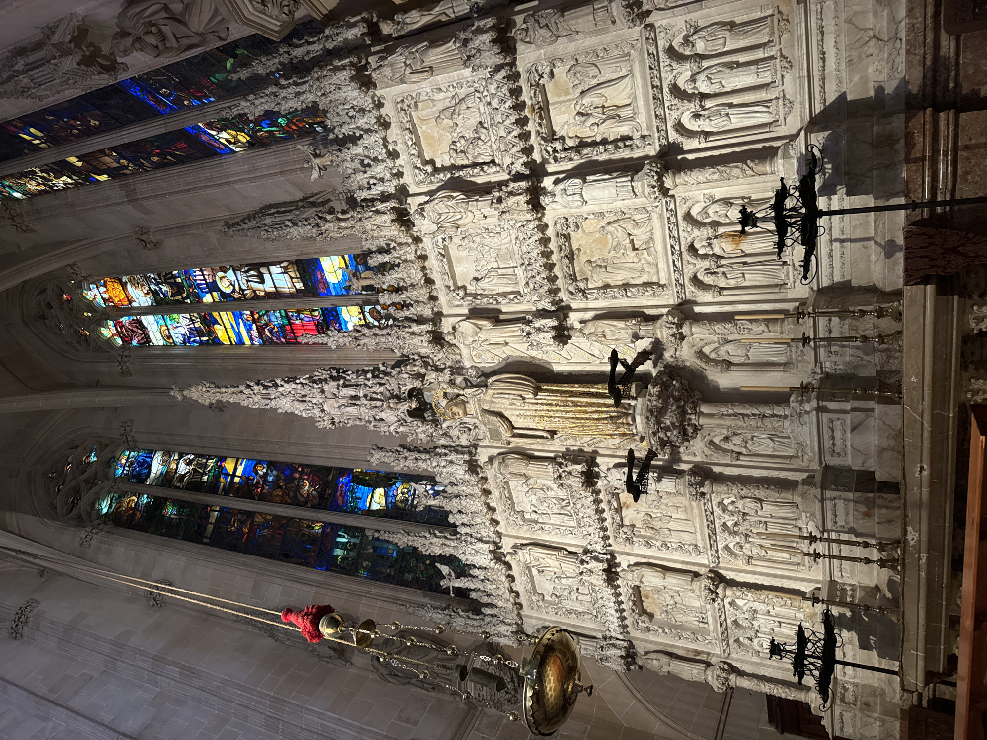 The image shows the interior of a cathedral, likely the Cathedral of Santa Maria of Palma, located in Palma de Mallorca, Spain. The scene is dominated by a large, ornate altar wall featuring intricate stone carvings and a central gilded statue of a saint. Above the altar wall are tall, narrow stained-glass windows with vibrant colors, depicting religious scenes. To the left, a hanging lamp with a red tassel and a brass bowl is suspended by a rope. The stone architecture of the cathedral, with its vaulted ceilings and imposing columns, is visible in the background. The lighting suggests an indoor, possibly dimly lit environment, with the stained glass windows providing colorful illumination. In the foreground, several candelabras and a portion of a wooden bench are visible, indicating this is an active place of worship. The overall impression is one of grandeur, religious devotion, and historical significance.