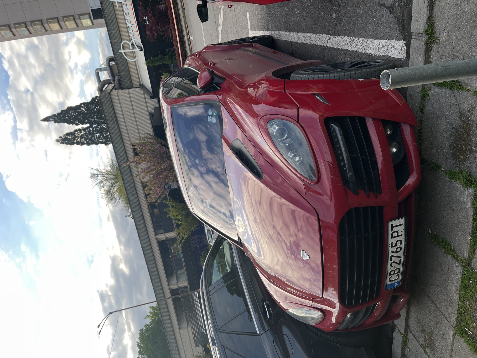 A custom red sports car, likely a tuned Porsche Cayenne, is parked on an asphalt street in Sofia, Bulgaria, captured during daytime under a partly cloudy sky. The car is positioned diagonally, showcasing its aggressive front design with a large black grille, circular headlights, and LED daytime running lights. Visible customizations include hood vents, a bespoke front bumper, and black multi-spoke wheels. A black stripe is present on the side of the car's red body.
The front license plate, "BG CB 2765 PT," confirms its Bulgarian registration, with "CB" denoting Sofia. The text "CARGOMOBILE.BG" is also present below the license plate. Reflections of the cloudy sky are visible on the car's glossy surface and windshield.
In the background, urban architecture is visible, including a modern building with multiple windows and a large white "B" shaped logo, along with another structure featuring horizontal paneling. Green trees and bushes with pink blossoms add natural elements to the scene, suggesting a spring or early summer season. A dark-colored vehicle, possibly an SUV, is partially visible parked next to the red car. A grey metal pole stands in the foreground to the right of the car's front. No people are visible, and the car appears to be stationary.