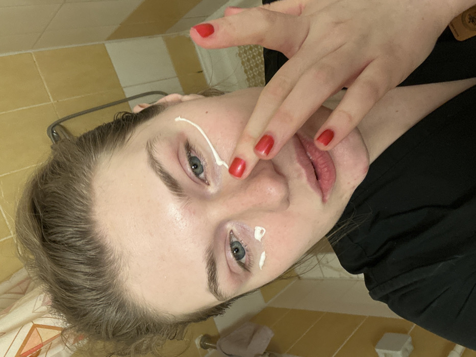A young woman's face is close to the camera, rotated slightly to the left. She has fair skin, light brown hair, and blue eyes. She's applying white cream or lotion to her face with her fingers, which have red nail polish. One dollop of cream is on her cheek near her eye, another below the other eye, and she's using her finger to spread cream near her forehead. She's wearing a black top. The background includes a tiled wall and floor, suggesting a bathroom.