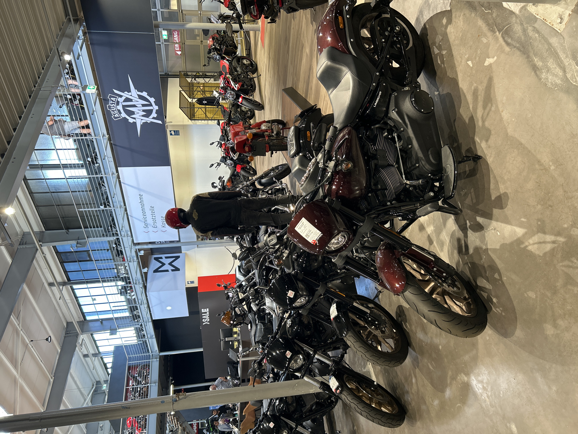 The image shows a motorcycle exhibition in Bernhausen, Germany. Numerous motorcycles of various brands are on display, with a focus on dark-colored models. A person wearing a red helmet and dark clothing stands among the motorcycles. Banners with motorcycle brand logos are suspended from the ceiling, including the MV Agusta logo. Signs indicating "Serviceannahme" (service reception), "Ersatzteile" (spare parts), and "Kasse" (cashier) are visible. The environment is a large, open space with a concrete floor and a high ceiling with metal beams. A "SALE" sign is also apparent. The motorcycles are arranged in rows, and the overall impression is that of a commercial exhibition or dealership.