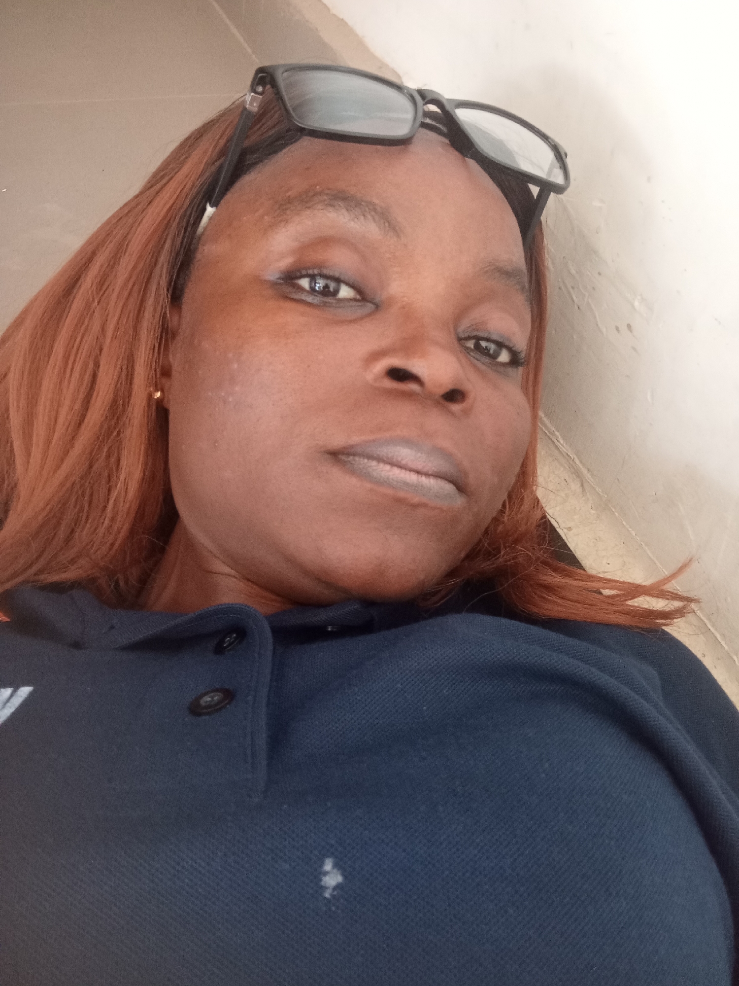 The image is a close-up, head-and-shoulders portrait of a Black woman with reddish-brown hair. She is wearing glasses on top of her head and a dark blue collared shirt with two dark buttons visible on the placket. Her expression is neutral. The background is a light-colored, textured wall. There is a small, light-colored smudge or stain on the shirt. The lighting suggests it is daytime, and the overall scene appears to be indoors or in a shaded outdoor area. There are no discernible location cues or text in the image, apart from the possible logo on the shirt which is not clear enough to read.