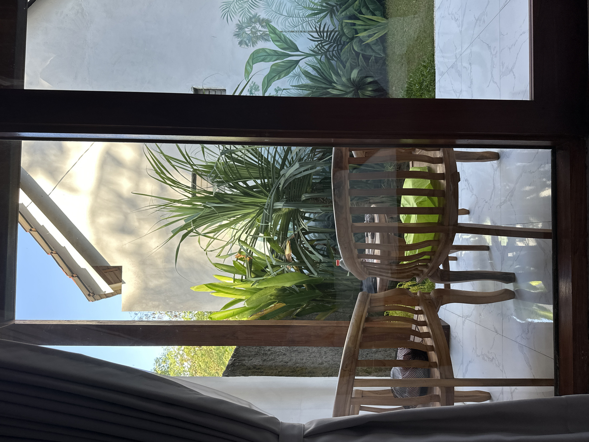 From an indoor perspective, the view through a window frame in Nusa Penida, Indonesia, encompasses an outdoor area. The foreground features wooden chairs, with one draped with a light green cloth. Beyond the chairs, the view transitions to tropical foliage, including palm trees. The backdrop includes a building facade and a wall adorned with a tropical plant mural. The foreground curtain is visible. The tiling is white with grey marbling.