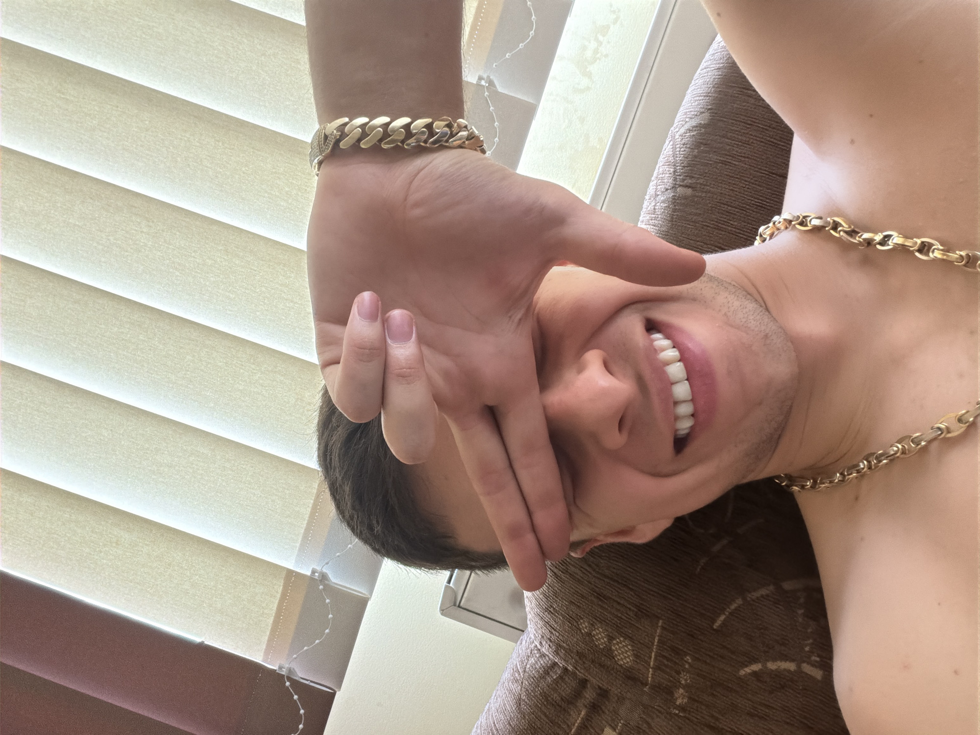 A man is positioned upside down in front of brown and beige blinds. He is wearing a gold chain necklace and a gold bracelet on his left wrist. His left hand is obscuring part of his face, and he is smiling. He is sitting on a brown patterned couch. The location is Unknown, Unknown.