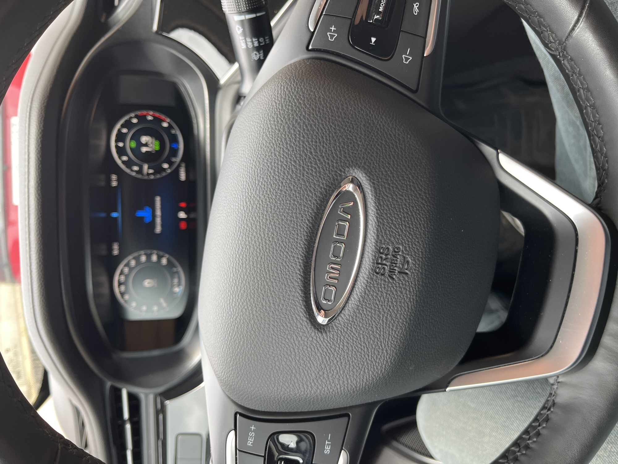 The image shows the interior of a car, focusing on the steering wheel and dashboard. The steering wheel has a black leather cover with a silver "OMODA" logo in the center, accompanied by "SRS AIRBAG" inscription. Control buttons are visible on both sides of the steering wheel. The dashboard features a digital display with gauges, including a circular indicator displaying a number "13". The surrounding environment is partially visible, suggesting the car is likely stationary. The location in the city of Sochi, Russia, is not discernible from the visual elements of the image.