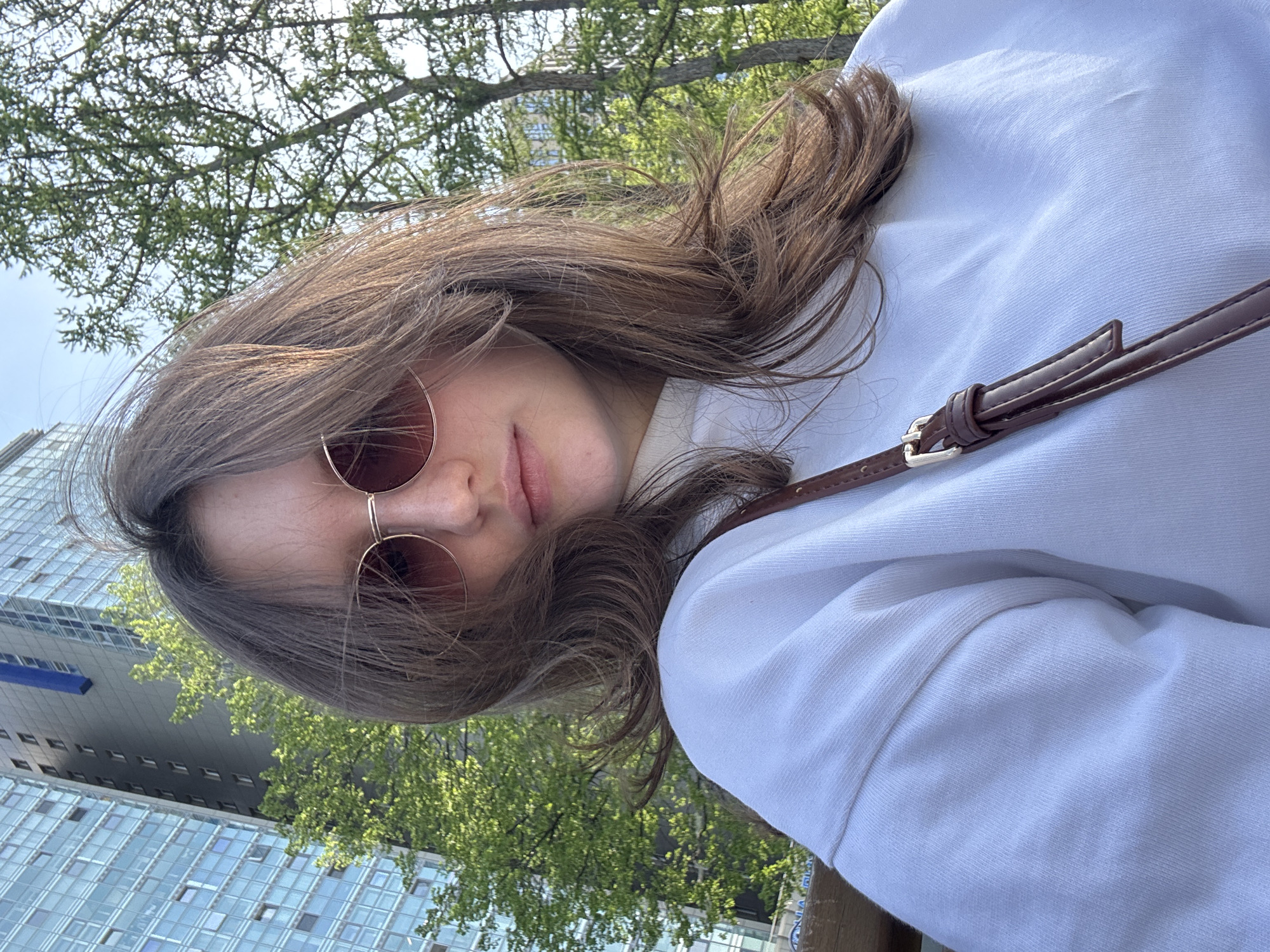 A medium shot presents a woman with shoulder-length, light brown hair and wearing rose-tinted sunglasses. She is turned slightly to her left. She's wearing a white sweatshirt and a brown, leather-like purse strap is visible across her body.
In the background, there's a building with a blue-grey facade and many windows. Above her, tree branches with green leaves are visible against a light sky.
The location is identified as Perm, Russia.