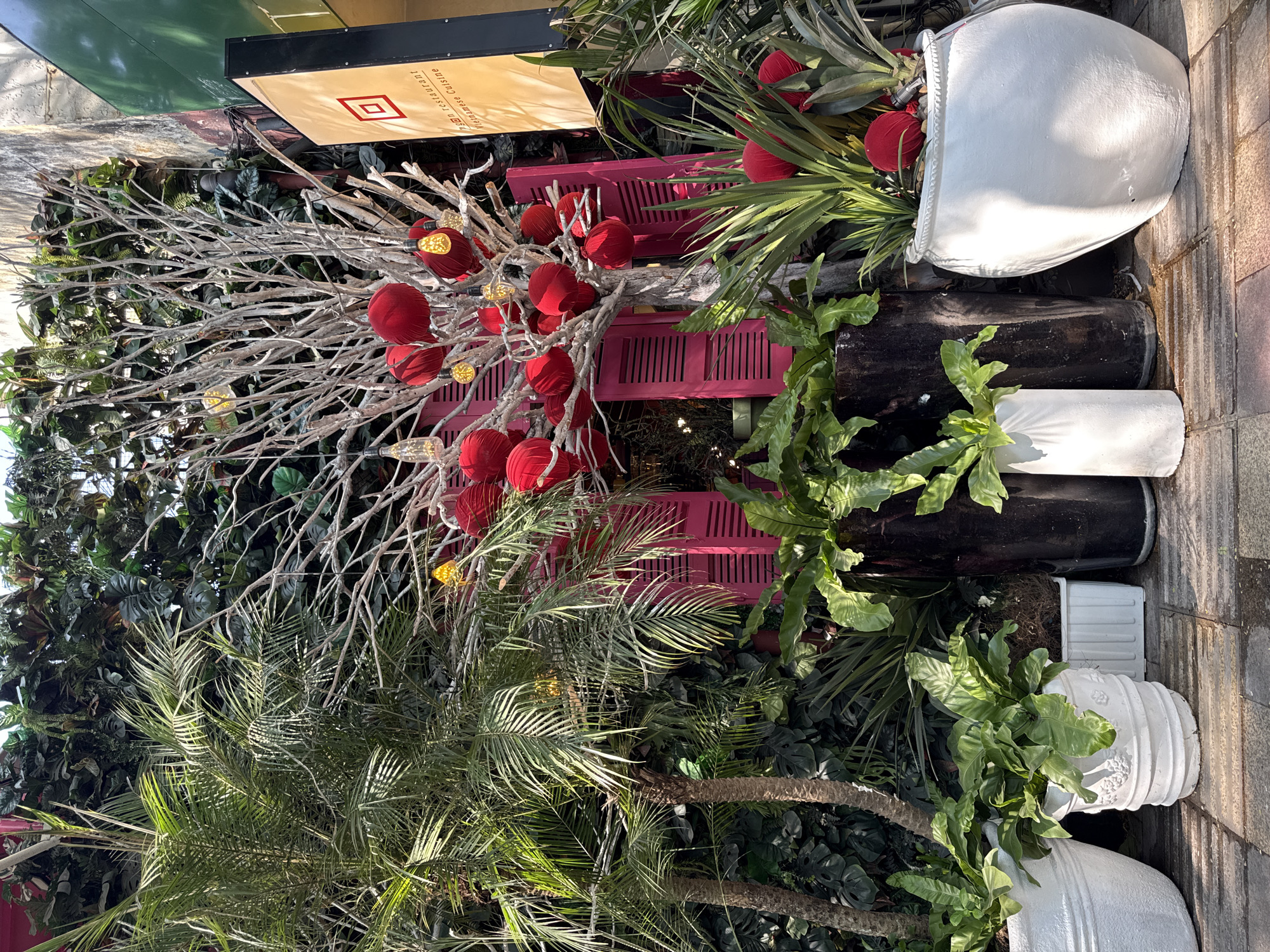 The image captures a vibrant street-side scene in Thủ Đức, Vietnam, featuring a decorative display that combines natural and artificial elements. A dense cluster of plants, including palms and ferns, form a lush backdrop. In front, a painted white tree adorned with red spherical ornaments and small lights stands beside a hot pink trellis-like structure. Various planters, including large white ceramic pots and black cylindrical containers, house the vegetation. A sign partially visible at the top indicates a restaurant. The composition suggests an outdoor setting, likely a restaurant or business facade designed to attract attention.