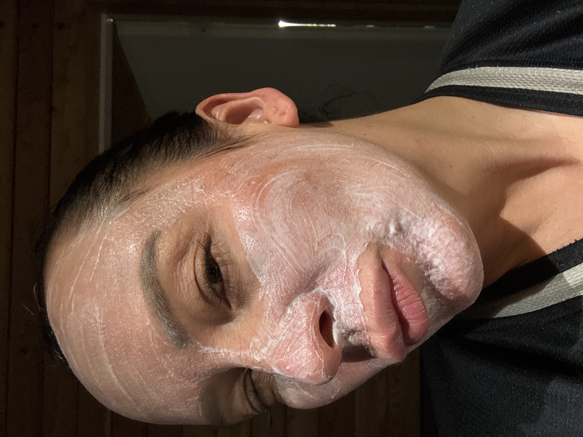 A close-up, tilted view shows a person's face covered in a white, creamy substance, likely a face mask. The person has dark hair and an ear is visible. The person is wearing a black and white striped shirt. The background is partially visible and appears to be a dark wooden door and frame. There is no information available to determine that this is in the city of Chaikovsky, Russia.