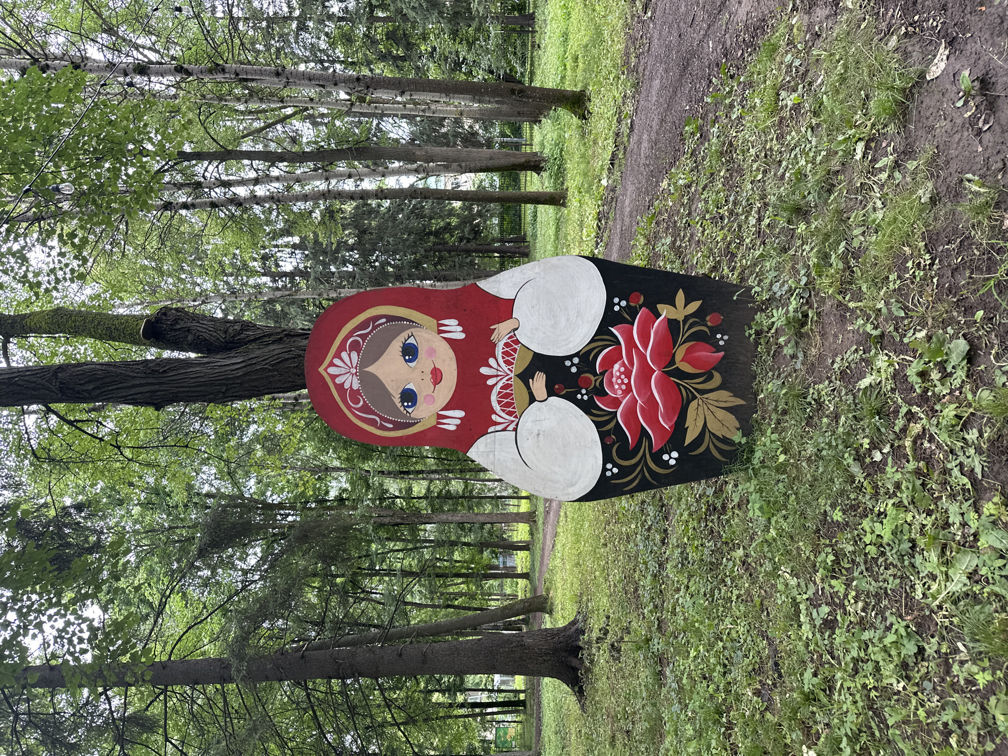 A large, painted Matryoshka doll stands upright in a park in Izhevsk, Russia. The doll is painted with a red headscarf, blue eyes, and a floral design on its black body, featuring red flowers and gold accents. It is positioned in front of a large tree, with a dirt path visible to the right and a dense forest with tall trees and greenery surrounding the area. The ground is covered in grass and fallen leaves. The weather appears overcast.