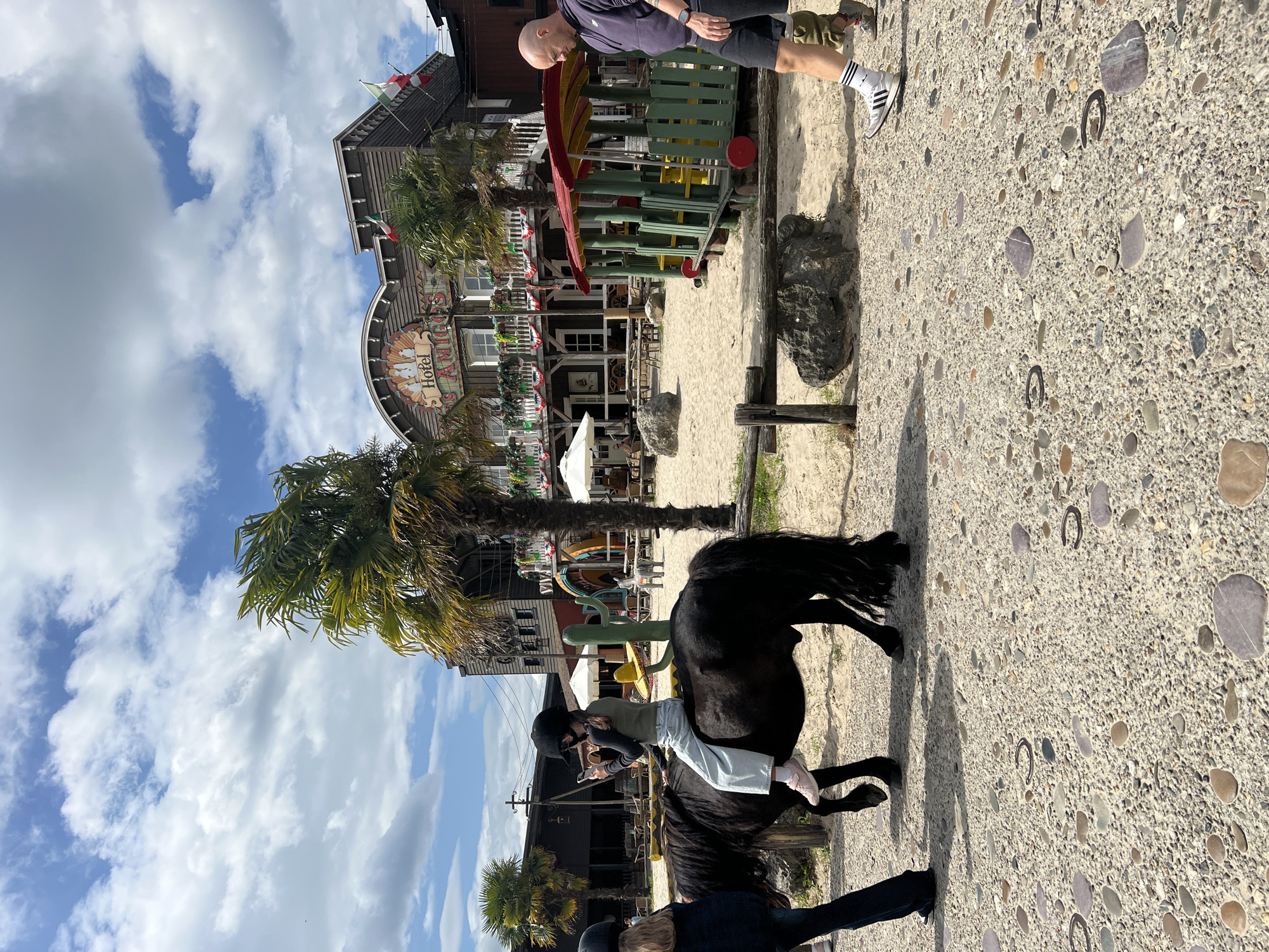 The image depicts an outdoor scene in Collendoorn, Netherlands, likely a theme park or entertainment venue, given the rustic Western-style architecture of the "Hotel Los Amigos" building in the background. In the foreground, a young person wearing a helmet, dark long-sleeved shirt, and light blue jeans is riding a small black pony. The rider is holding a smartphone in their left hand. To the left of the pony, another person, mostly out of frame, is walking. On the right, a bald man wearing a dark hoodie and black shorts with white socks and sneakers is walking away from the camera. The ground is a mix of sand and textured pavement with visible pebbles. Behind the pony, a wooden fence separates the sandy area from the paved walkway. The background features palm trees, white umbrellas, tables and chairs, and a colorful playground structure resembling a train. The sky is partly cloudy.