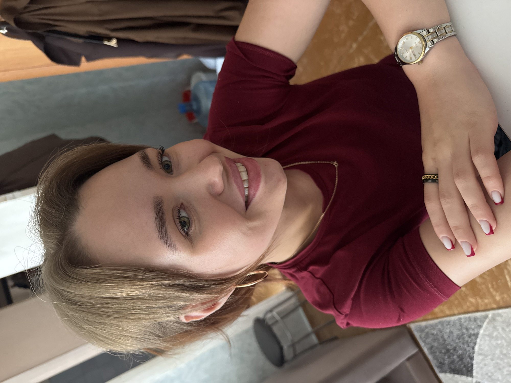 A young woman with blonde hair and green eyes is captured in a close-up, upper body shot. She is wearing a maroon t-shirt and a gold watch on her left wrist. She also wears two rings on her right ring finger, one black and gold, and another gold band. Her nails are painted with a French manicure style, with white tips and dark red bases. She has a gold chain necklace on. The woman is smiling broadly, with her head tilted slightly to her left.
The setting appears to be indoors, with a wooden surface visible in the background, possibly a table or desk. A bottle of water is also visible in the background. The lighting suggests daytime. The overall mood is cheerful and relaxed, conveyed by the woman's smile.
The image is oriented vertically, with the woman's head towards the top left of the frame. The provided crops highlight different aspects of the image, including close-ups of her face, hand with the watch and rings, and her hair.
No visible text is present in the image.
The location context is Chaikovsky, Russia.