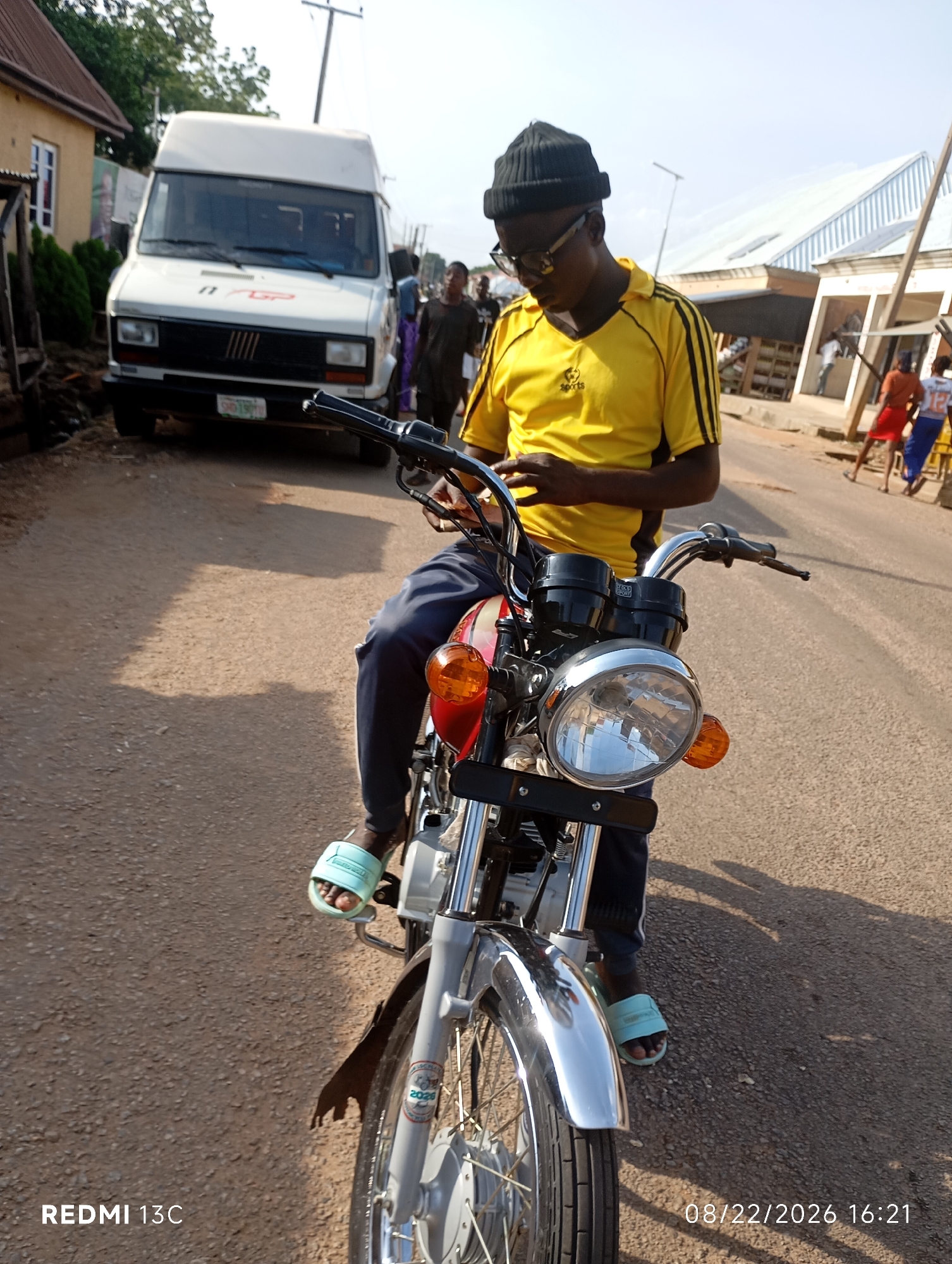 In Shendam, Nigeria, a person wearing a black beanie, glasses, and a yellow t-shirt with black stripes is sitting on a red and silver motorcycle. They are looking down at a phone. The motorcycle has a prominent chrome headlight. In the background, a white van is parked, and several other people are visible walking or standing. The scene is set on a paved road, with buildings and trees lining the street. The sky is bright and clear, suggesting daylight. The timestamp in the lower right corner indicates the image was taken on August 22, 2026, at 4:21 PM.