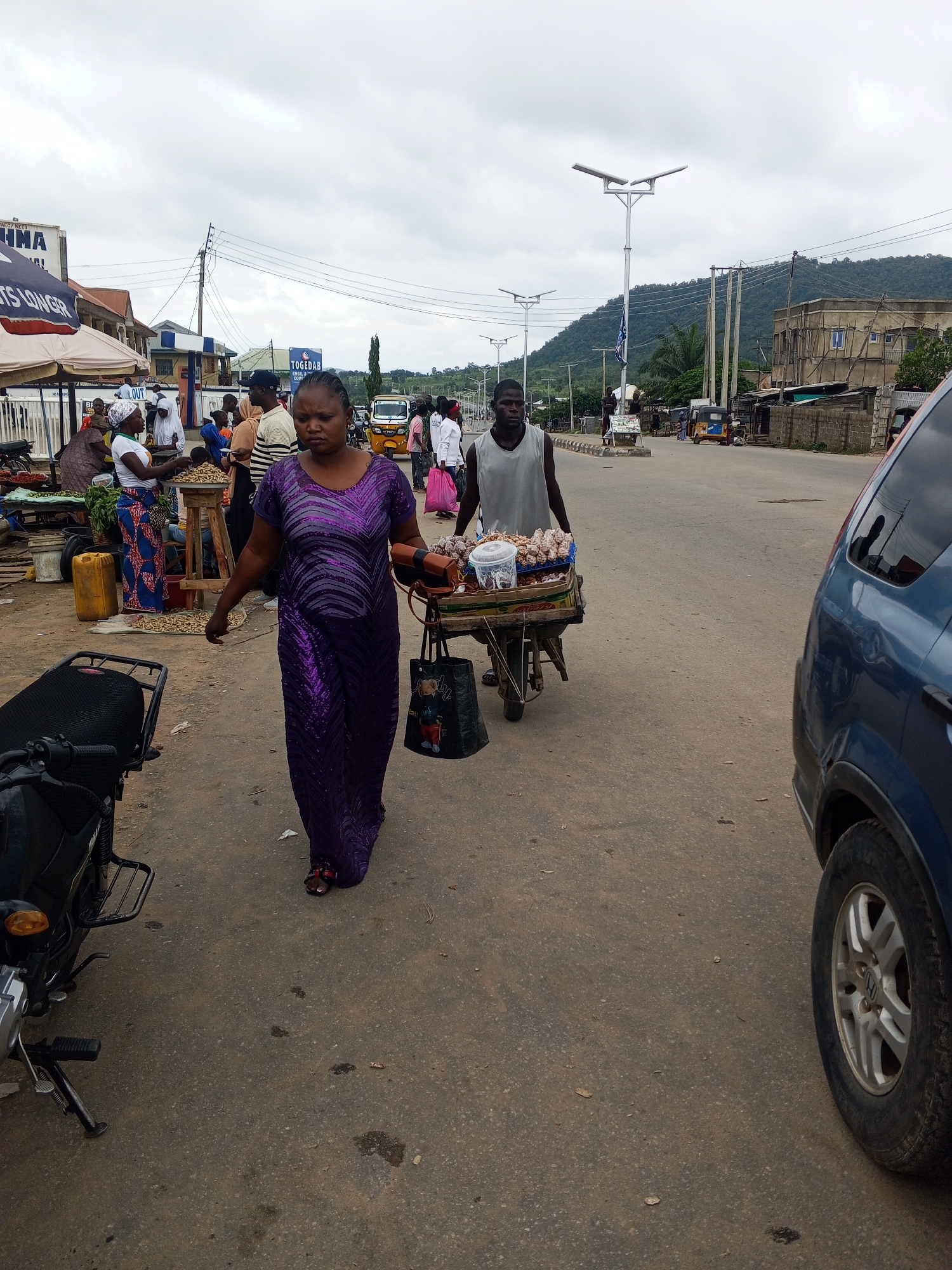 This image depicts a street scene in Zango-ndaji, Nigeria, under an overcast sky. In the foreground, a woman in a shimmering purple dress walks towards the right, carrying a small brown bag in her left hand and a larger black bag with a teddy bear design in her right. To her left, a portion of a parked motorcycle is visible, with its black seat and a silver handlebar. Behind the woman, a man wearing a light gray tank top pushes a cart laden with goods, including a clear plastic container and what appear to be skewers of food.
Further back, the street is lined with various stalls and buildings. A woman in a blue and red patterned skirt stands behind a stall displaying peanuts. Other individuals are seen interacting near the stalls, some wearing headscarves. Several vehicles are present on the road, including a yellow tricycle (keke napep) and a blue SUV partially visible on the right side of the frame. Utility poles with wires run along the street, and in the background, a hilly landscape with dense vegetation is visible beneath the cloudy sky. Signage for businesses such as "TOGEDAB" and "IMA" are discernable on buildings. The overall environment suggests a bustling market or roadside commercial area.