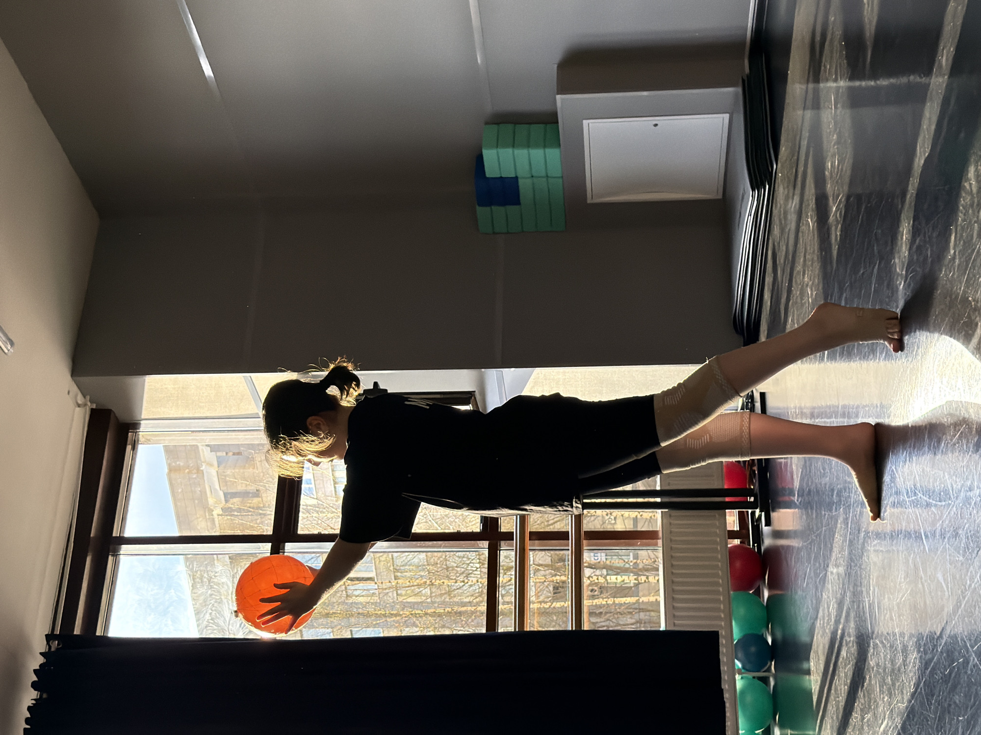 A person is depicted indoors, seemingly in a gymnasium or dance studio, performing a balance exercise near a window. The person is wearing dark clothing, with what appears to be support wraps around their lower legs. They are positioned on their stomach, balancing on an apparatus while holding an orange ball with their hand. Outside the window, buildings and bare trees are visible. Based on the architecture visible through the window, the location is likely Saint Petersburg, Russia. The room contains exercise equipment like yoga blocks and stability balls. The floor is marked with scratches or lines. The overall lighting suggests it is daytime.