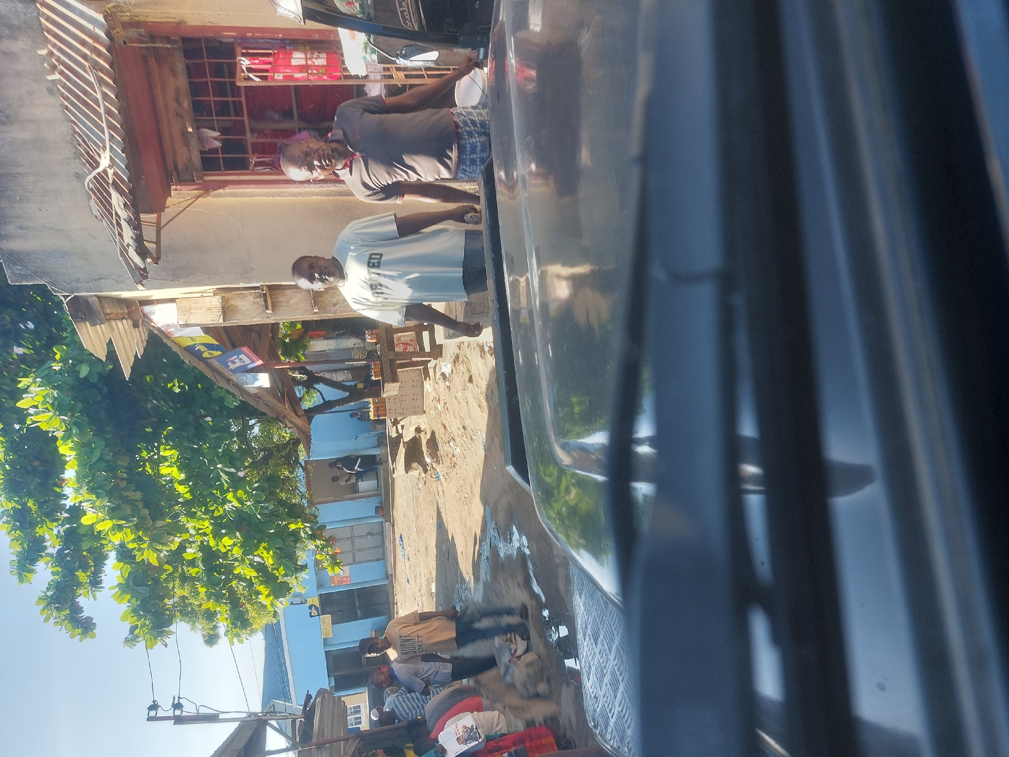 The image shows a street scene in Dar es-Salaam, Tanzania. Two men stand on the sidewalk in front of a building with a storefront. One man is wearing a light-colored t-shirt, and the other a darker t-shirt and patterned shorts. The street appears sandy and has puddles. Several other individuals and objects are visible in the background, along with buildings, trees, and power lines. The view is partially obscured by a large, dark object (likely part of a vehicle) in the foreground.