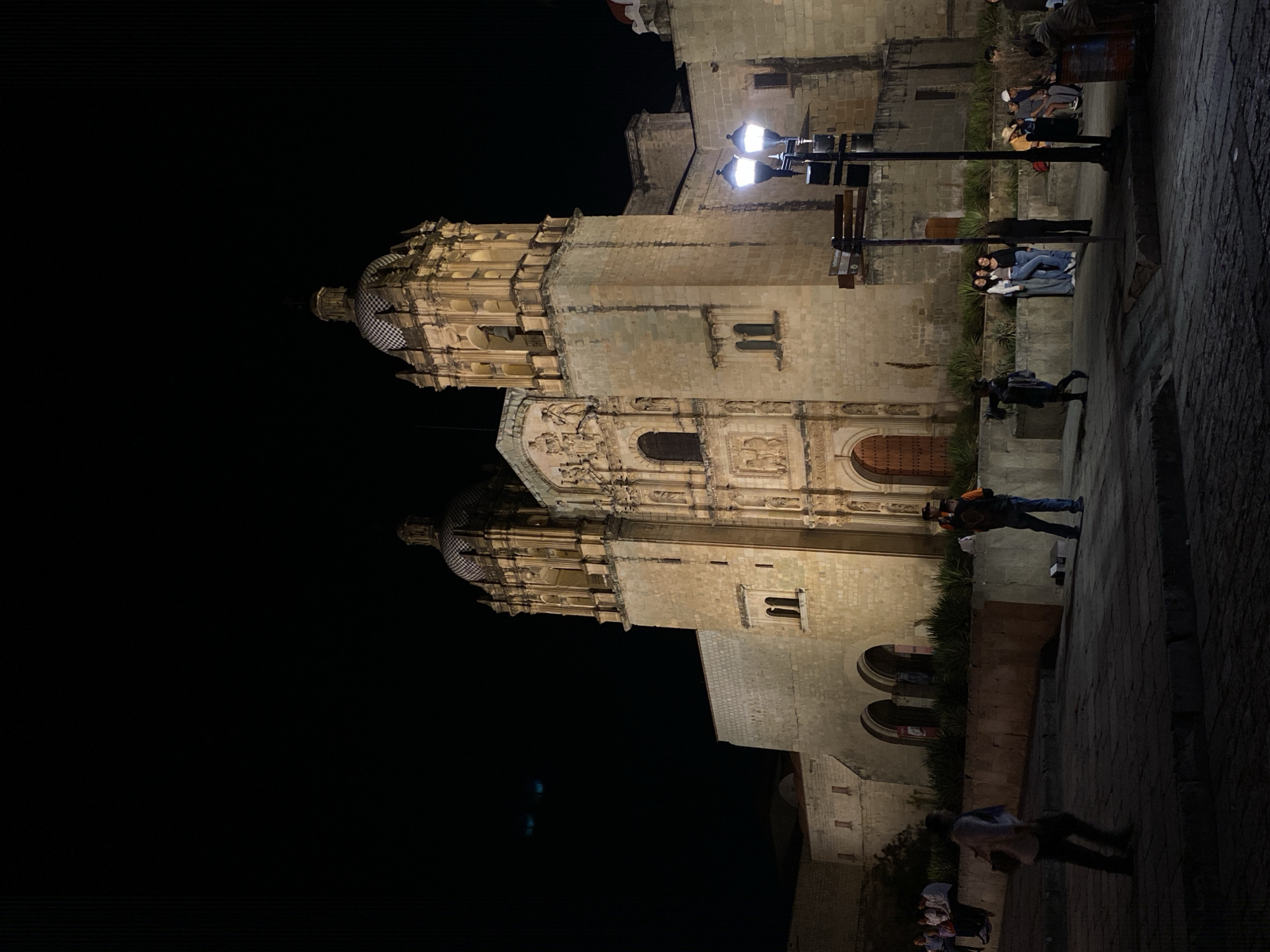 At night in Oaxaca City, Mexico, an angled, eye-level perspective captures the Santo Domingo de Guzmán church and the street activity in front. Constructed of light-colored stone, the church is partially illuminated. It features two bell towers topped with domes. People are visible on the street, including several sitting on a ledge. There is a lamppost next to the ledge. The sky is dark.